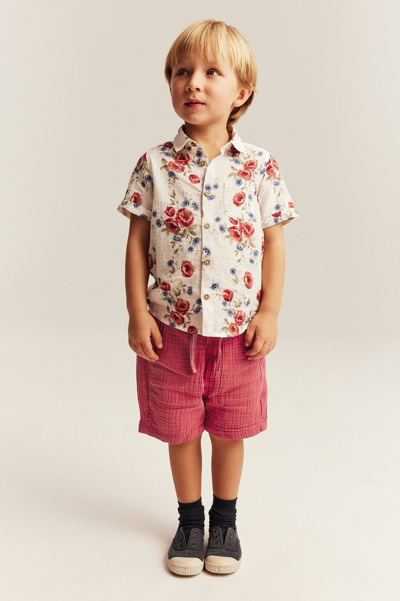 Framsida av ett barn i röda vävda bomullsshorts, vit blommig kortärmad skjorta, mörka strumpor och mörka tygskor.