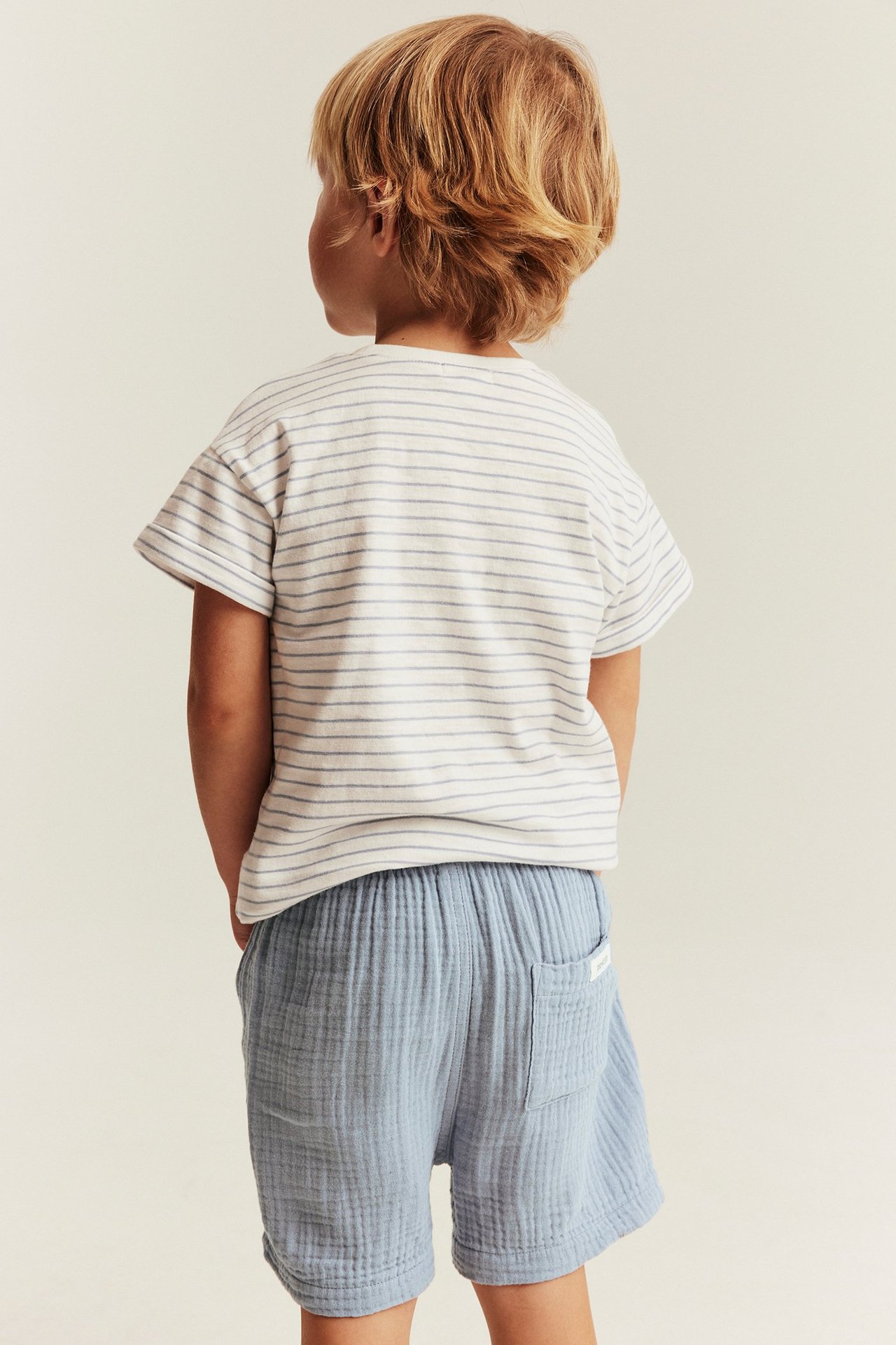 Widok z tyłu: Dziecko w niebieskich bawełnianych szortach z tylną kieszenią i pasiastej koszulce.