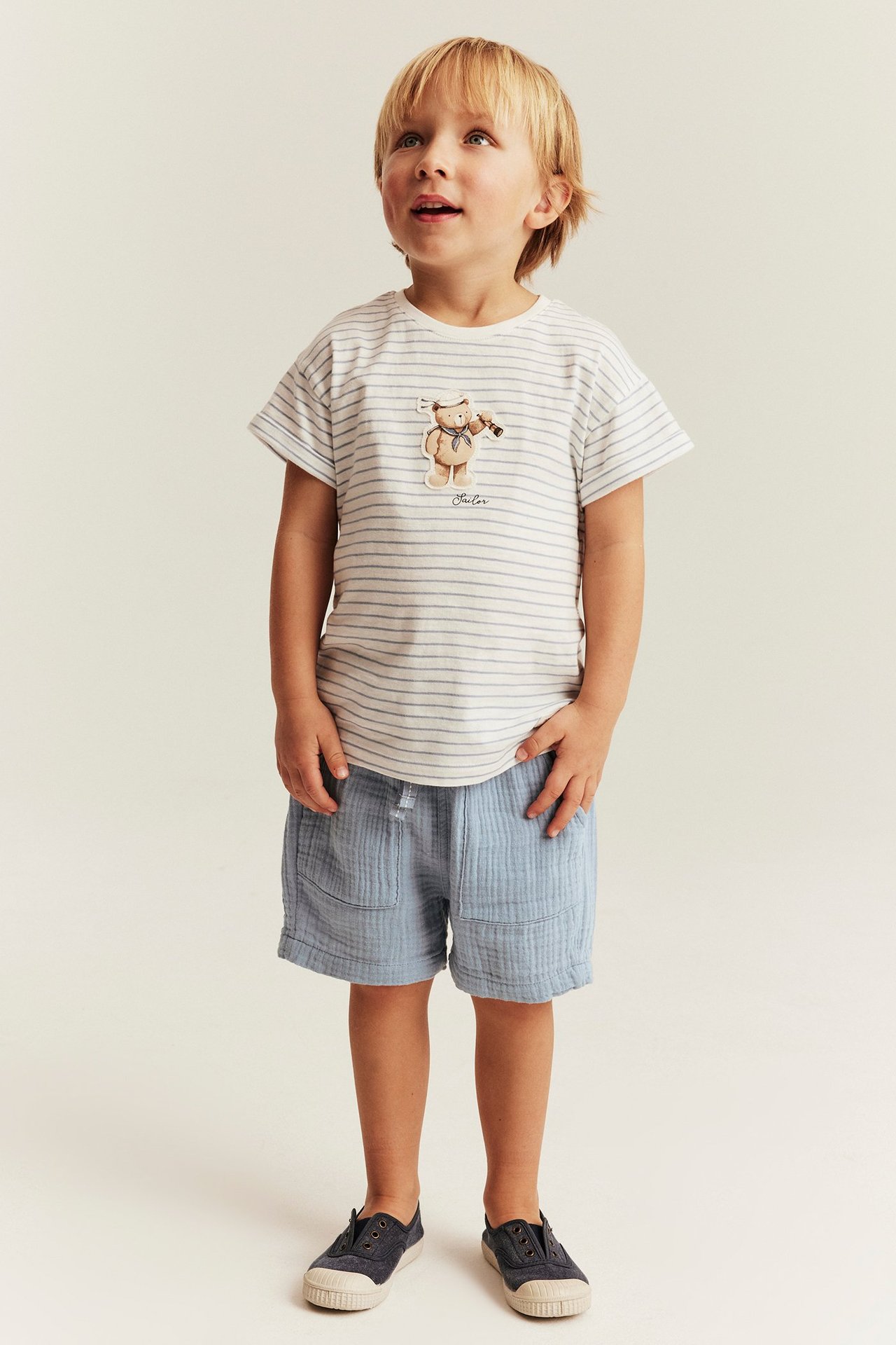 Frontvy av ett barn i blå vävda shorts med fickor, randig t-shirt och mörkblå tygskor.