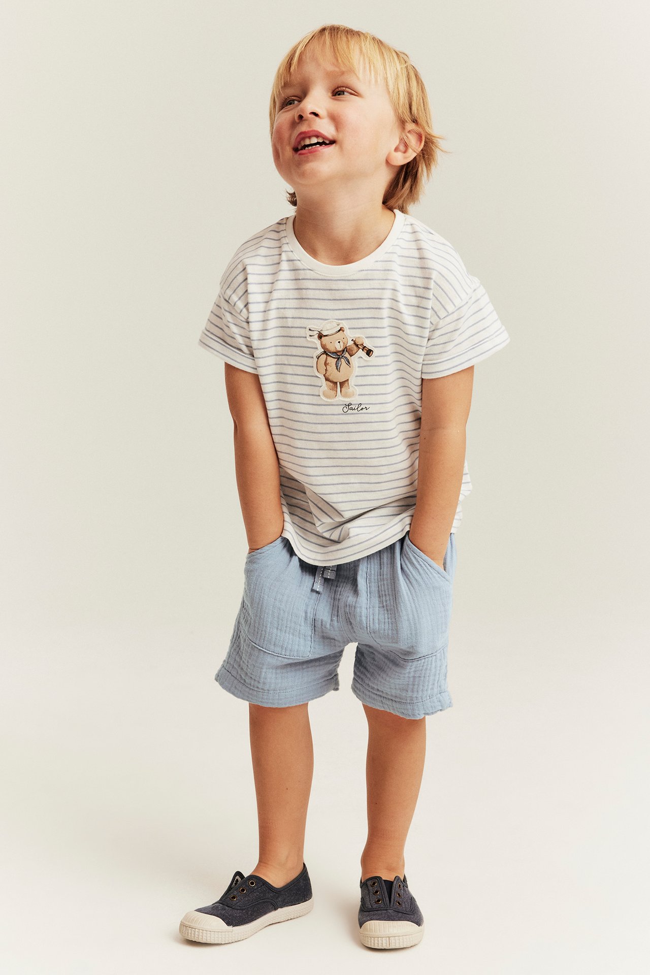 Edestä: poika sinisissä puuvillashortseissa taskuilla, raidallisessa t-paidassa, tummansinisissä kengissä.