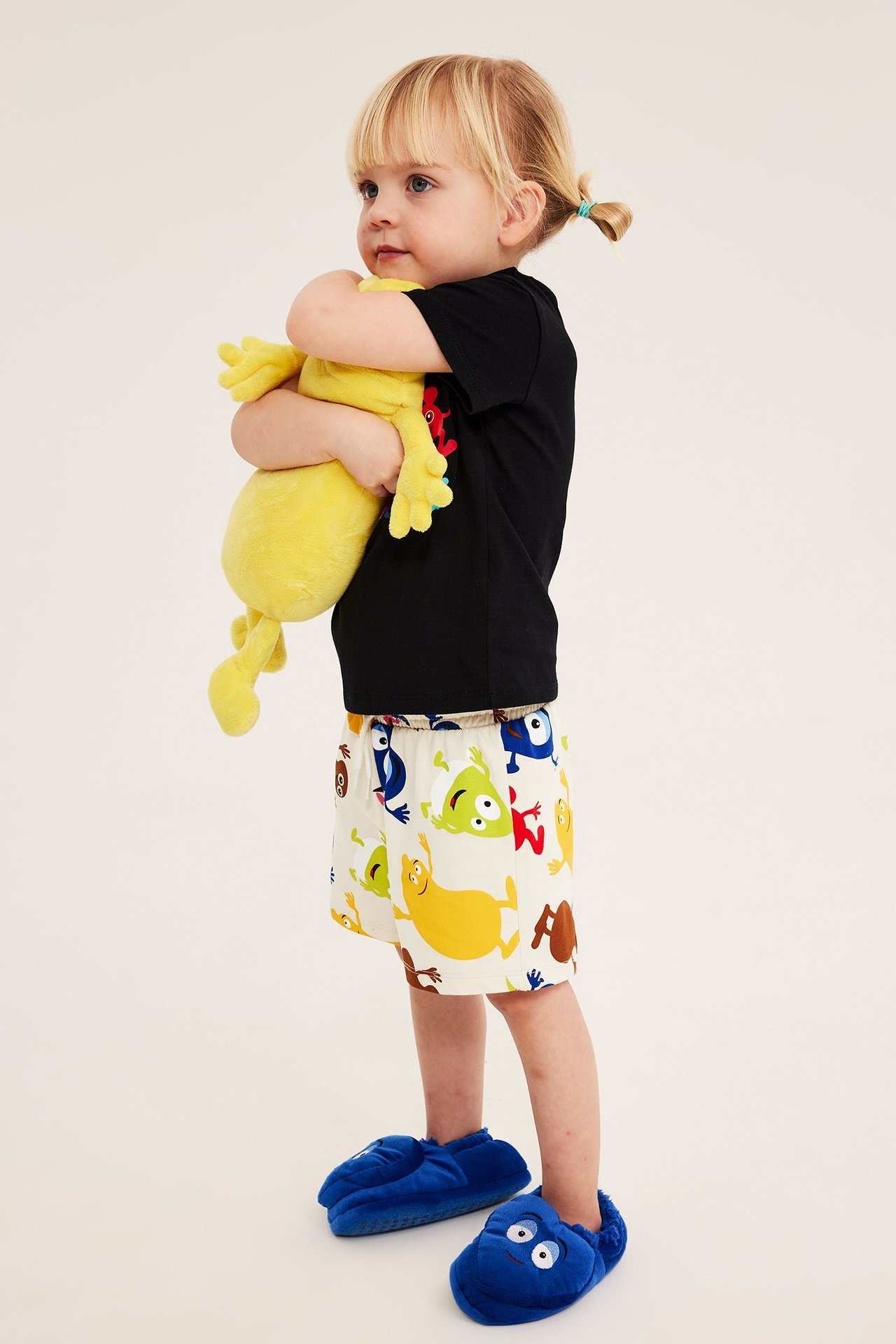 Widok z boku dziecka w jasnobeżowych wzorzystych szortach dresowych, czarnym t-shircie i niebieskich kapciach, trzymającego żółtą zabawkę.