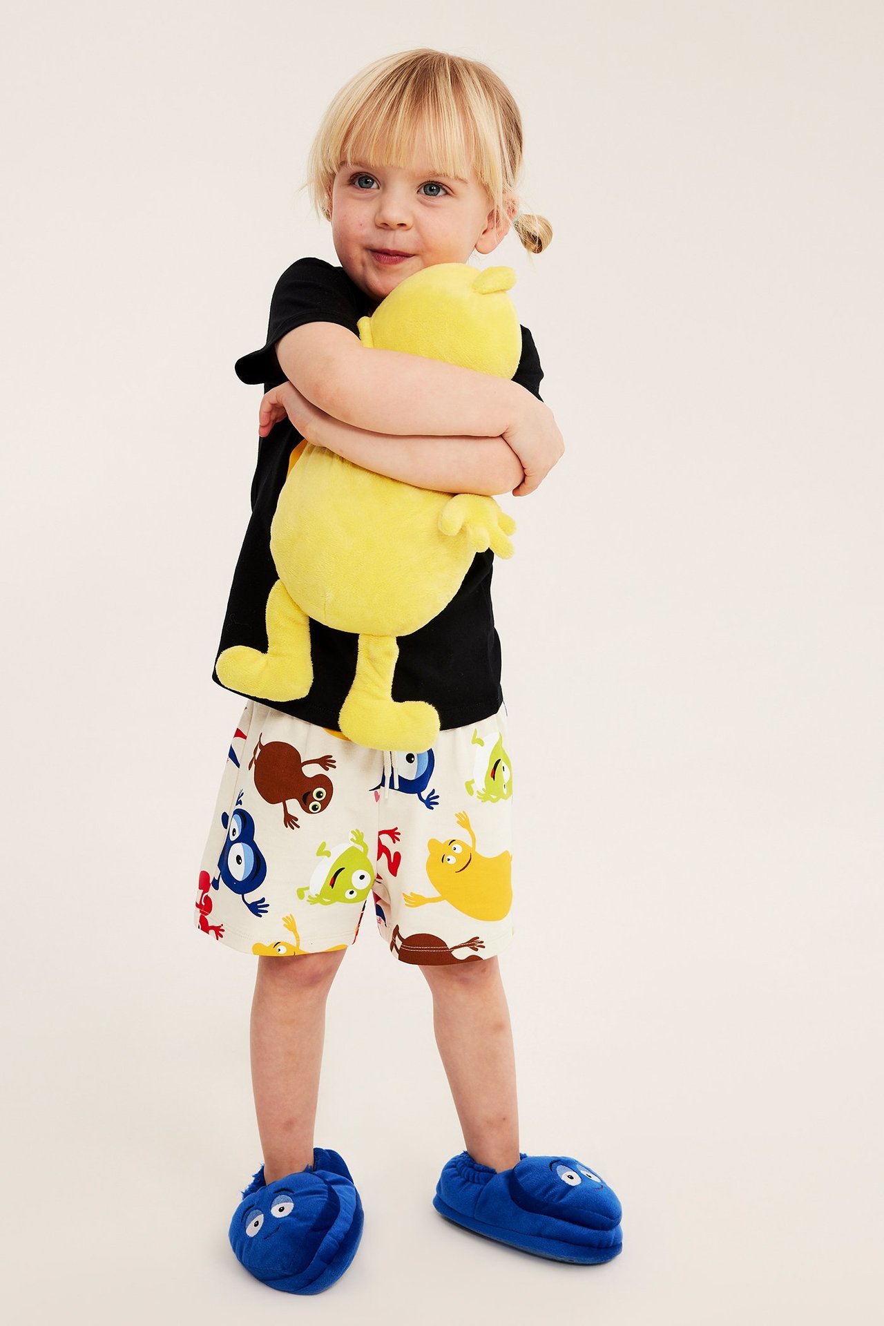 Widok z przodu: Dziecko w jasnobeżowych szortach Babblarna, czarnym t-shircie, niebieskich kapciach, trzyma żółtą pluszową zabawkę.