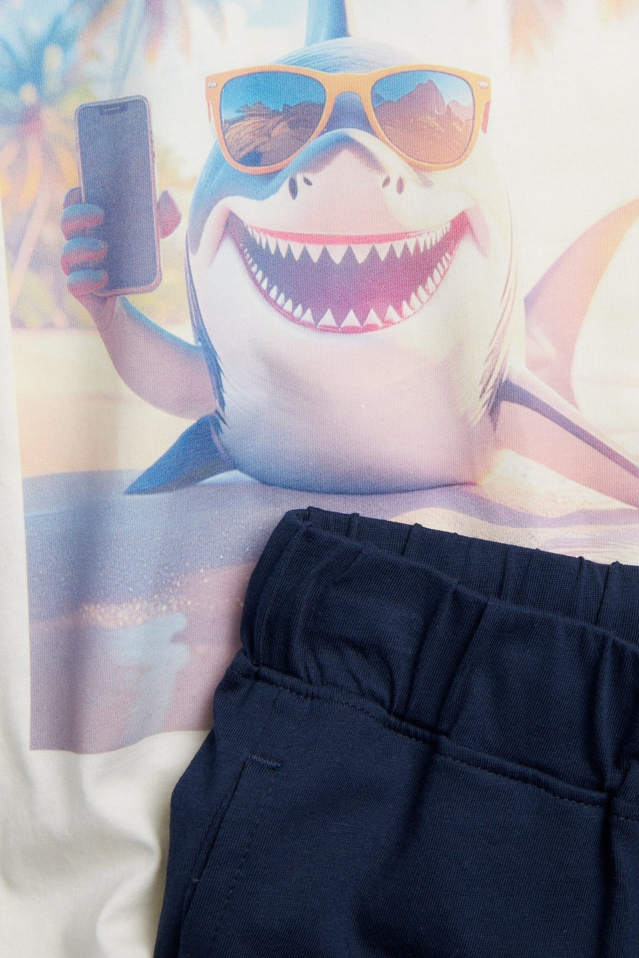 Lähikuva luonnonvalkoisesta pyjamapaidasta, jossa aurinkolasipäinen hai ja siniset shortsit.