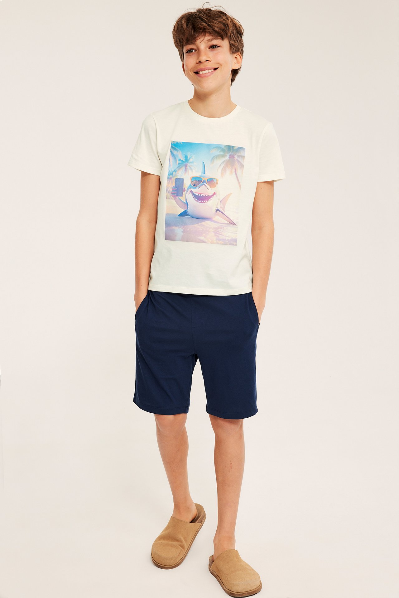 Widok z przodu: Dziecko w białawej, krótkiej piżamowej bluzce z rekinem, granatowych szortach i brązowych kapciach.