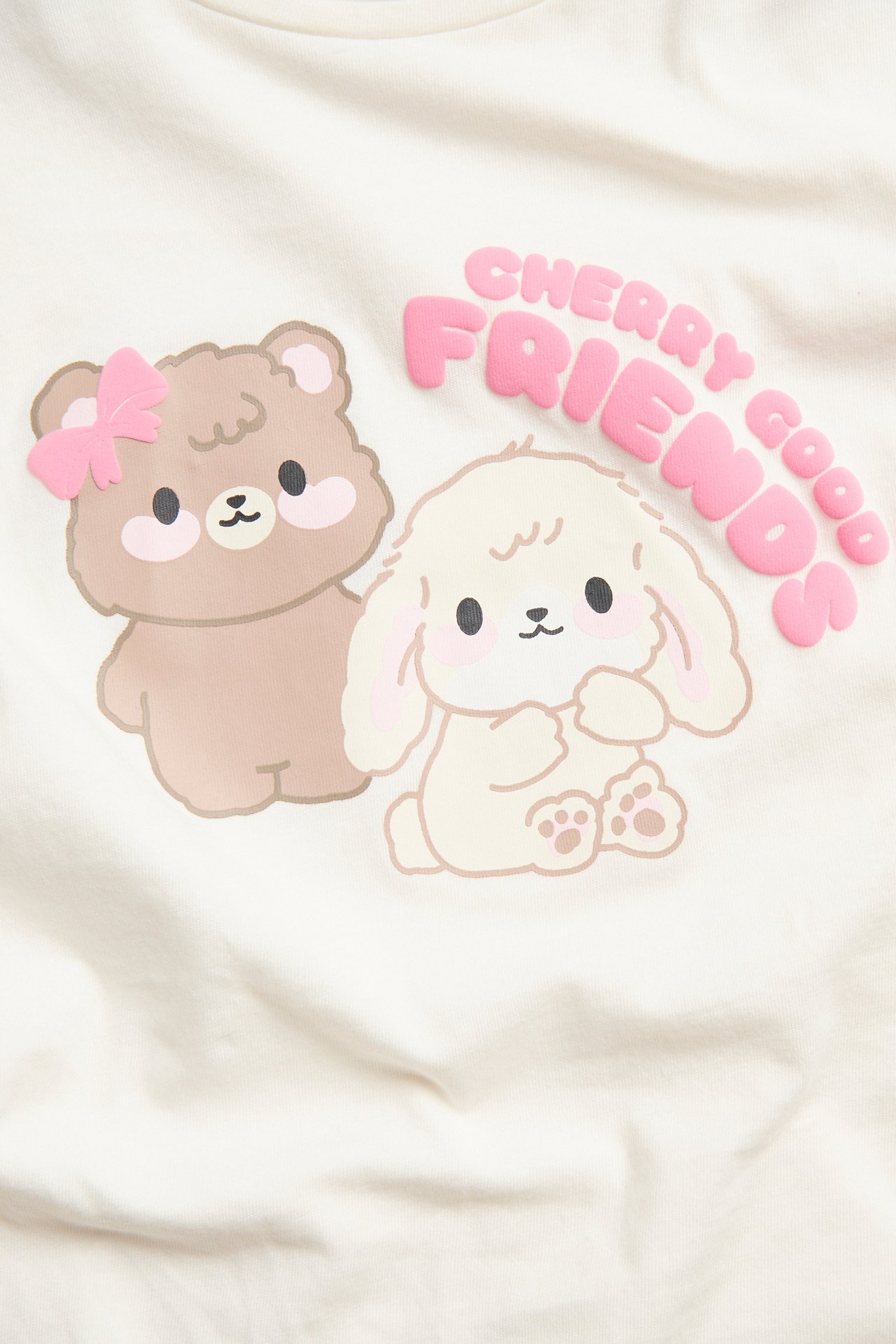 Lähikuva luonnonvalkoisesta puuvillaisesta pitkähihaisesta topista, jossa karhu, pupu ja 'CHERRY GOOD FRIENDS' -painatus.