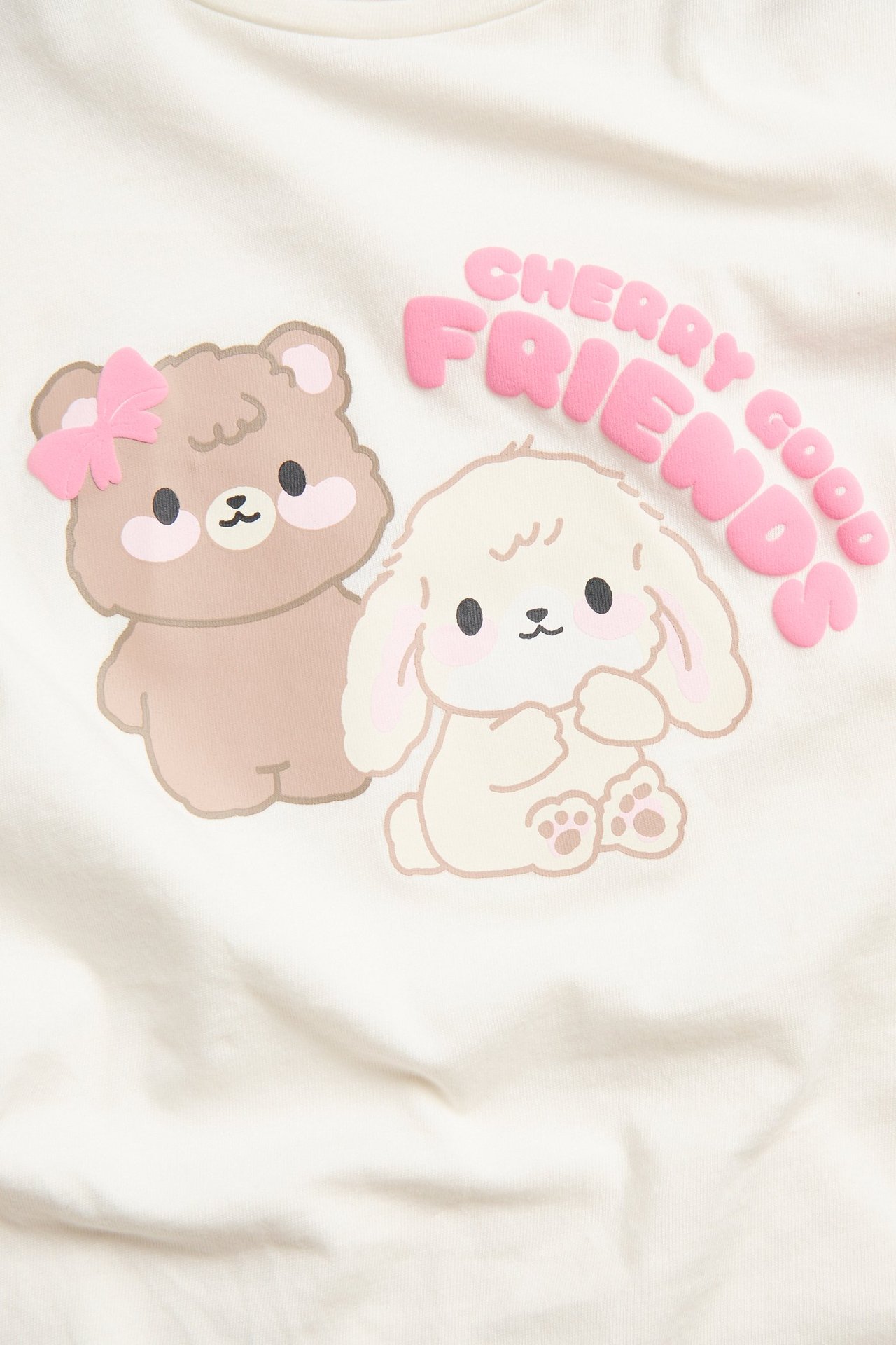 Zbliżenie na białawy bawełniany top z długim rękawem z nadrukiem misia, króliczka i 'CHERRY GOOD FRIENDS'.