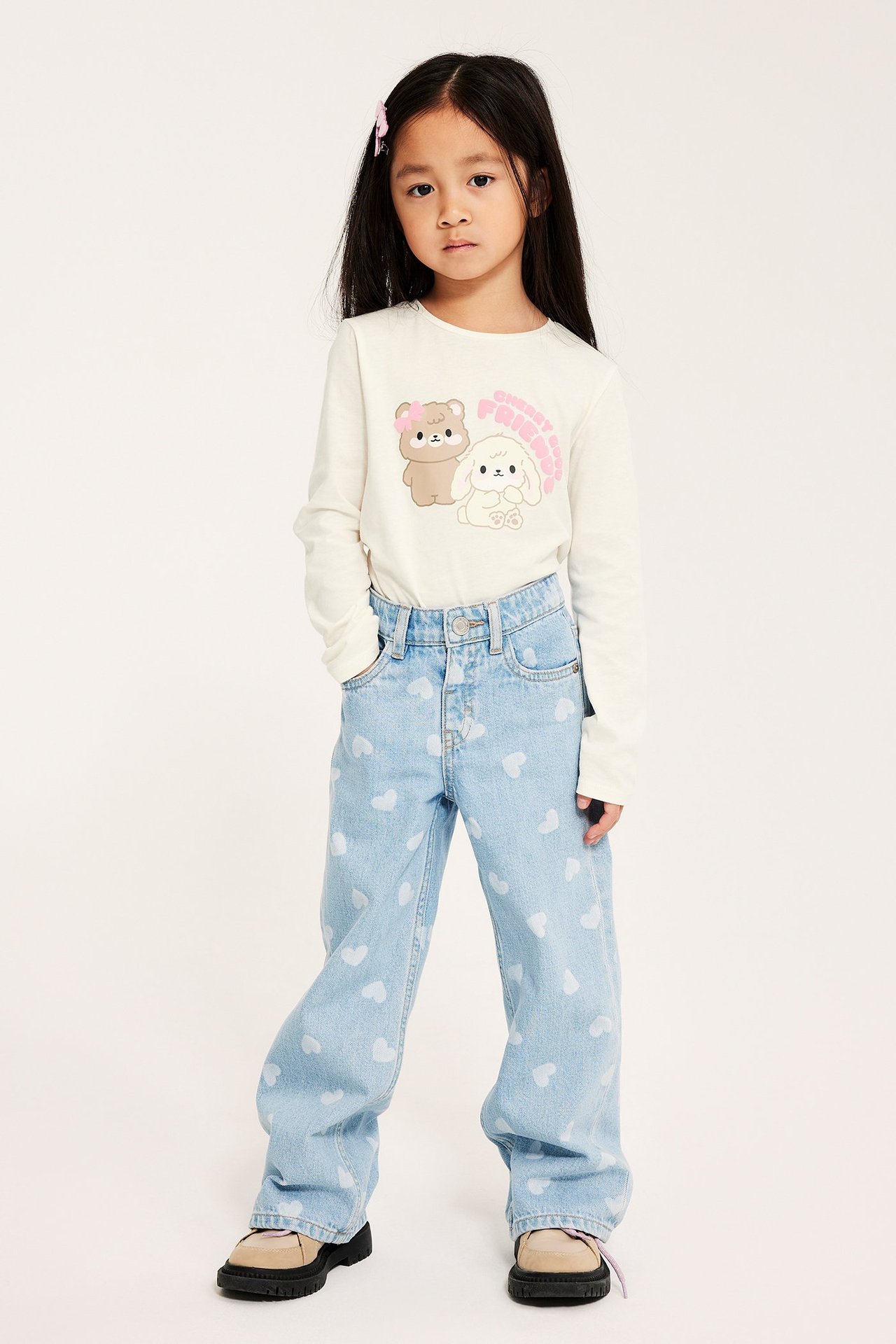 Frontbilde av et barn i en offwhite bomullstopp med lange ermer og dyreprint, lyseblå hjertejeans og loafers.