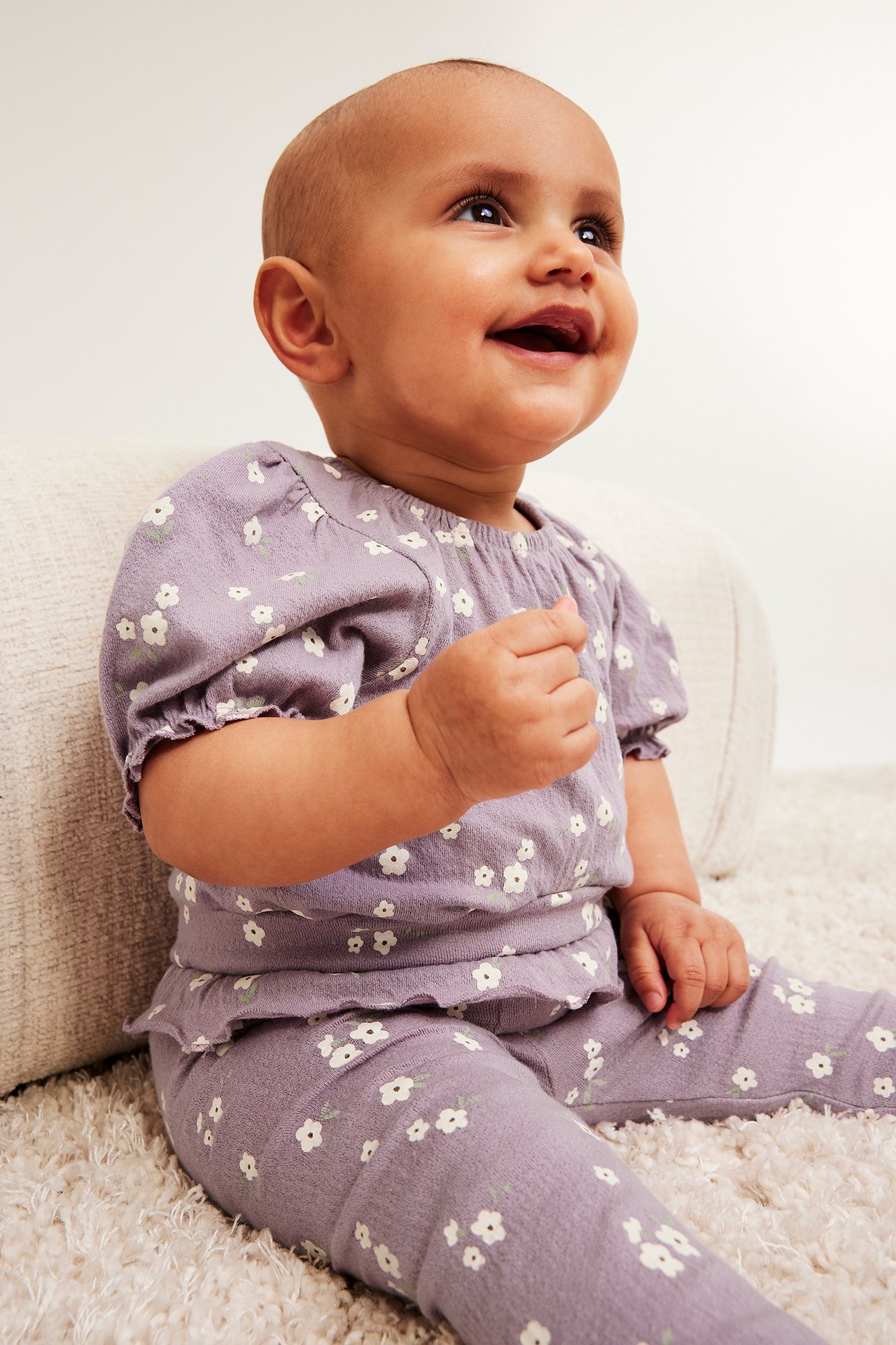 Frontvy av en baby i en lila blommig crepe kortärmad body med puffärmar och matchande leggings.