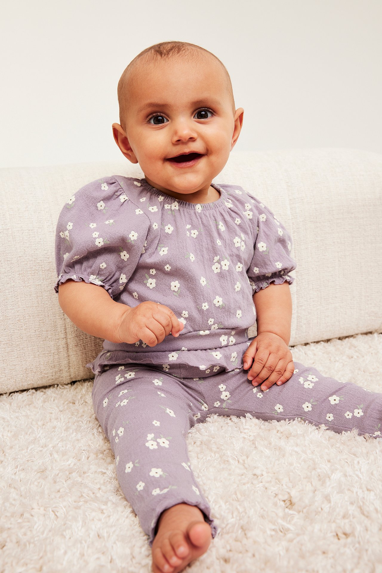 Widok z przodu: Niemowlę w liliowym body z krepy w kwiaty, z bufiastymi rękawami i pasującymi legginsami.