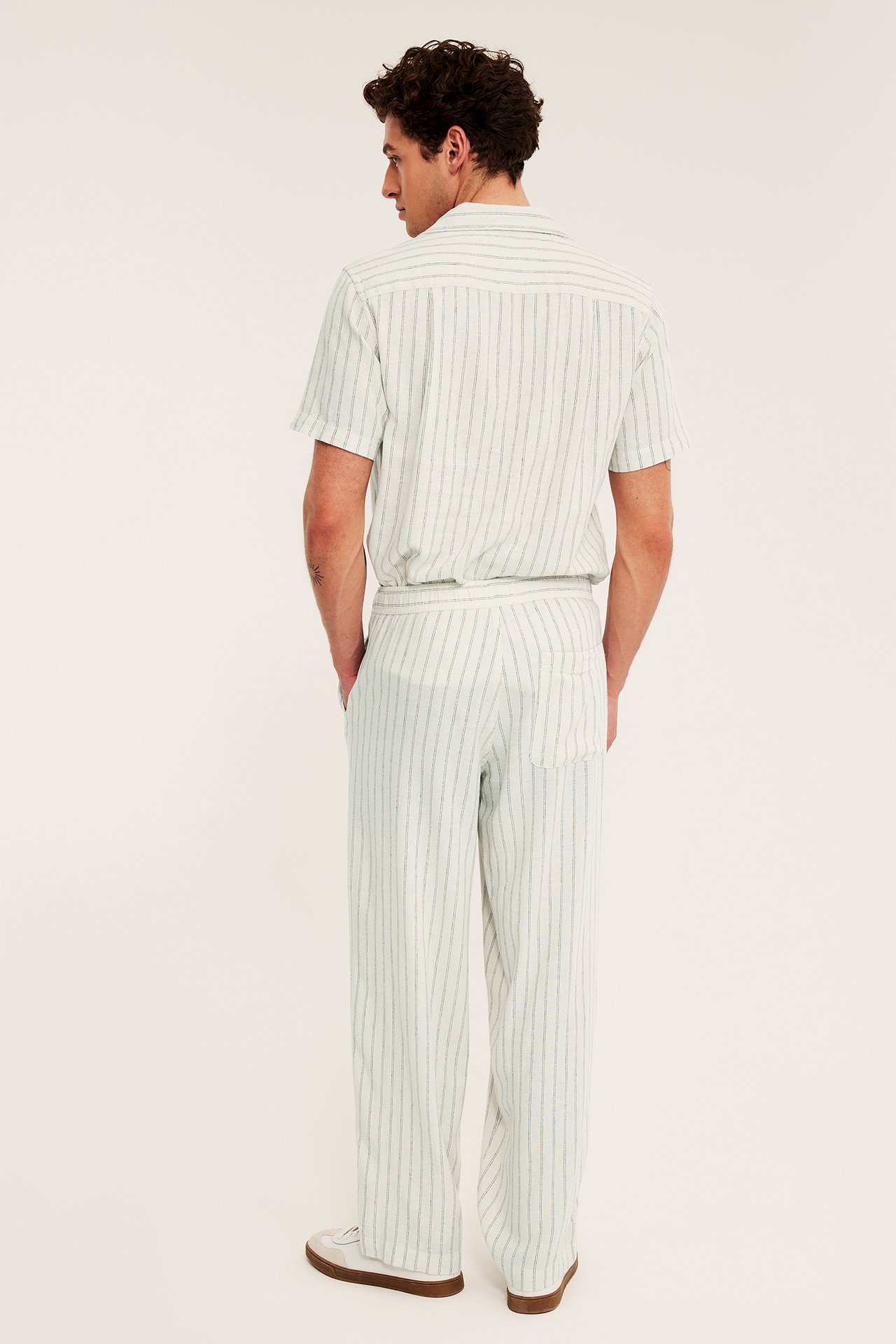 Mies selin päin raidallisissa beigeissä pellava-viskoosihousuissa taskuilla ja yhteensopivassa paidassa.