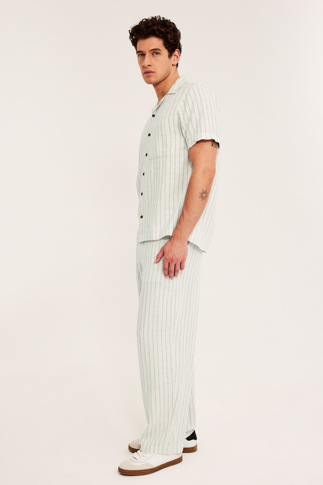 Sivukuva miehestä yllään beiget raidalliset pellava-viskoosihousut, yhteensopiva paita ja valkoiset tennarit.