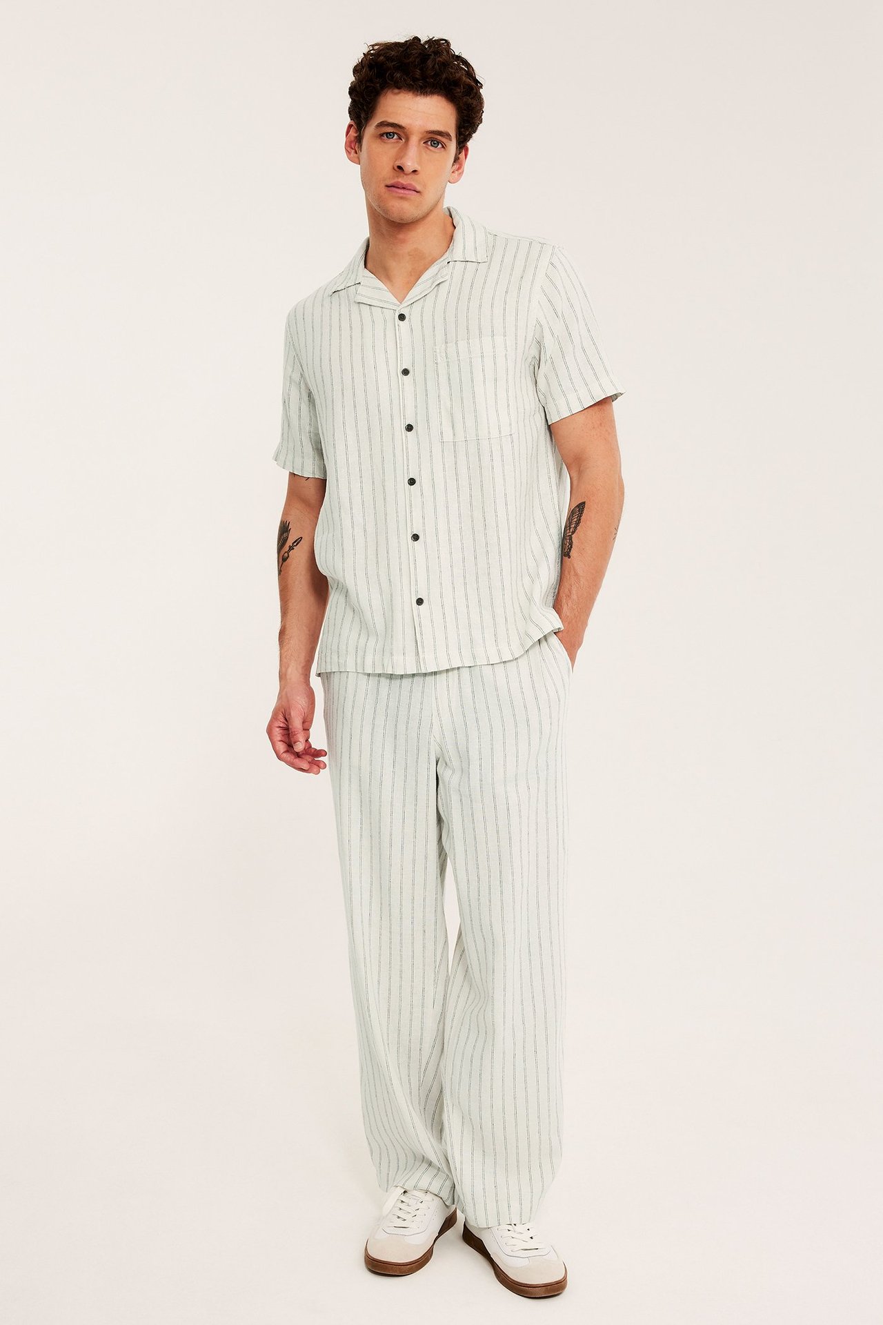 Etunäkymä: mies beigen raidallisissa pellavahousuissa, yhteensopivassa paidassa ja valkoisissa lenkkareissa.