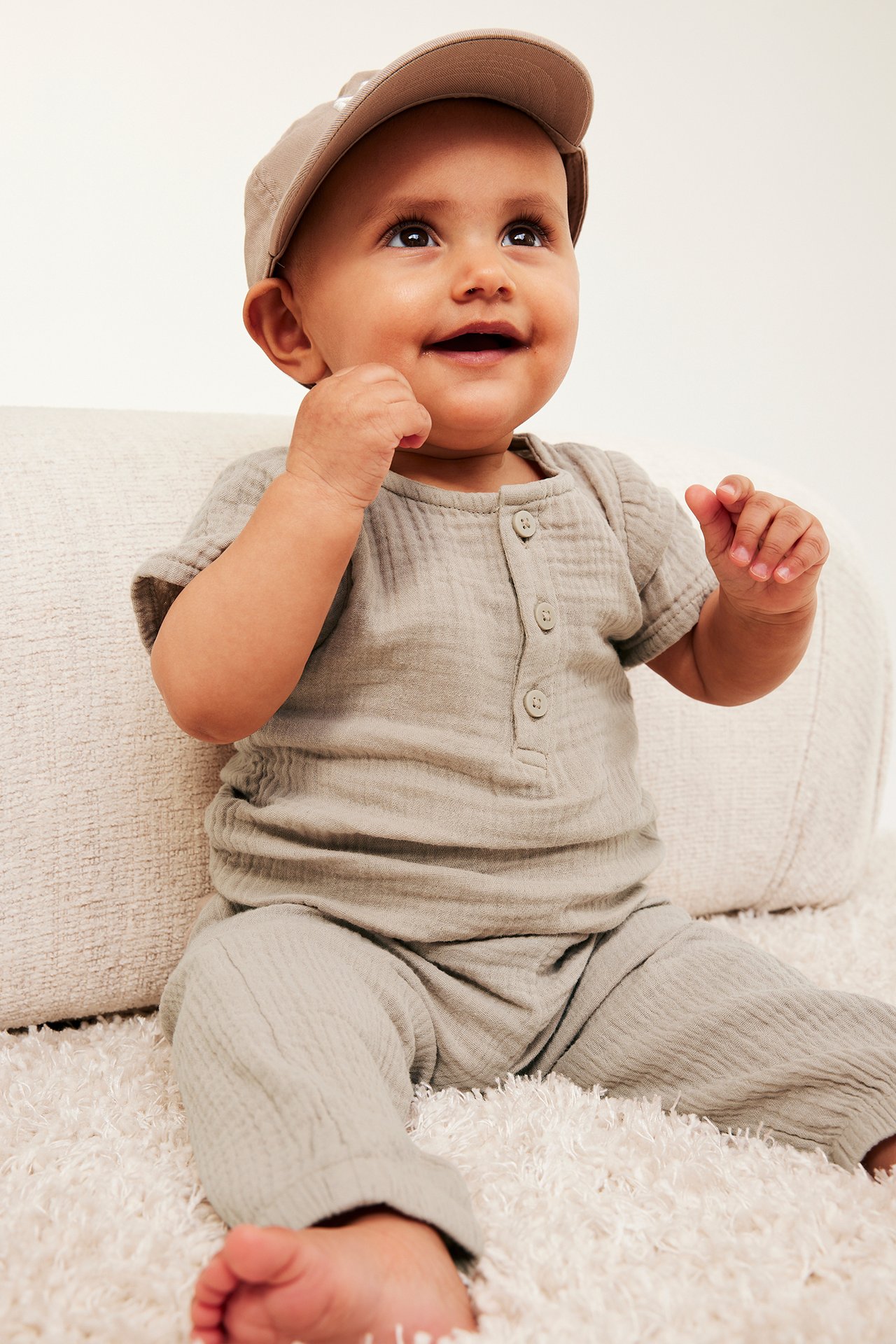 Frontvy: baby i beige kortärmad muslinbody med knappar, matchande byxor och en beige keps.