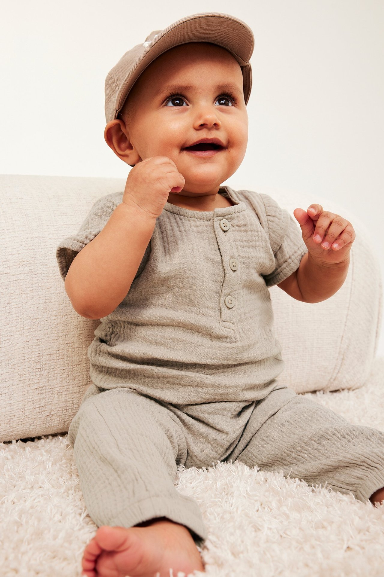 Edestä: vauva beigessä lyhythihaisessa musliinibodyssä napeilla, yhteensopivat housut ja beige lippalakki.