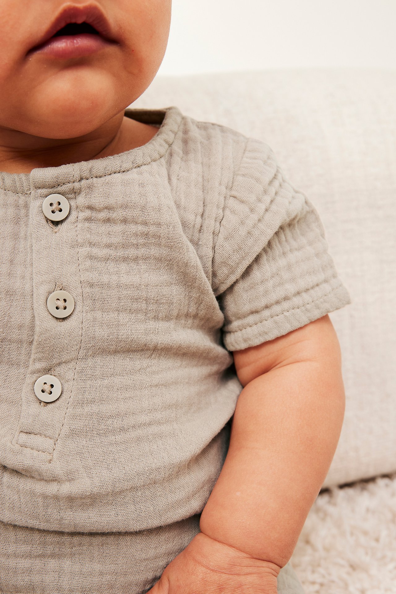 Närbild framifrån på en baby i beige kortärmad body i bomullsmuslin med knappar fram.