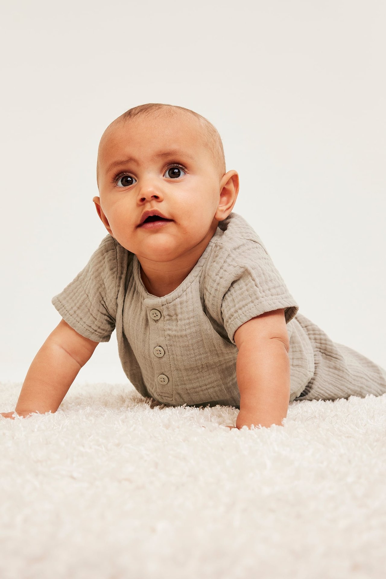 Frontvy av baby som kryper i en kortärmad beige muslinbody med knappar fram på en vit matta.