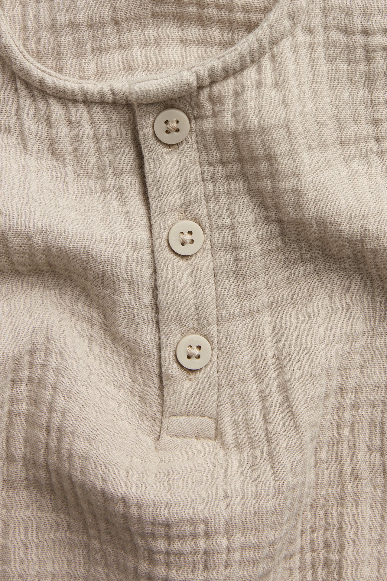 Nærbilde forfra av en beige babybody i bomullsmuslin med tre lyse knapper.