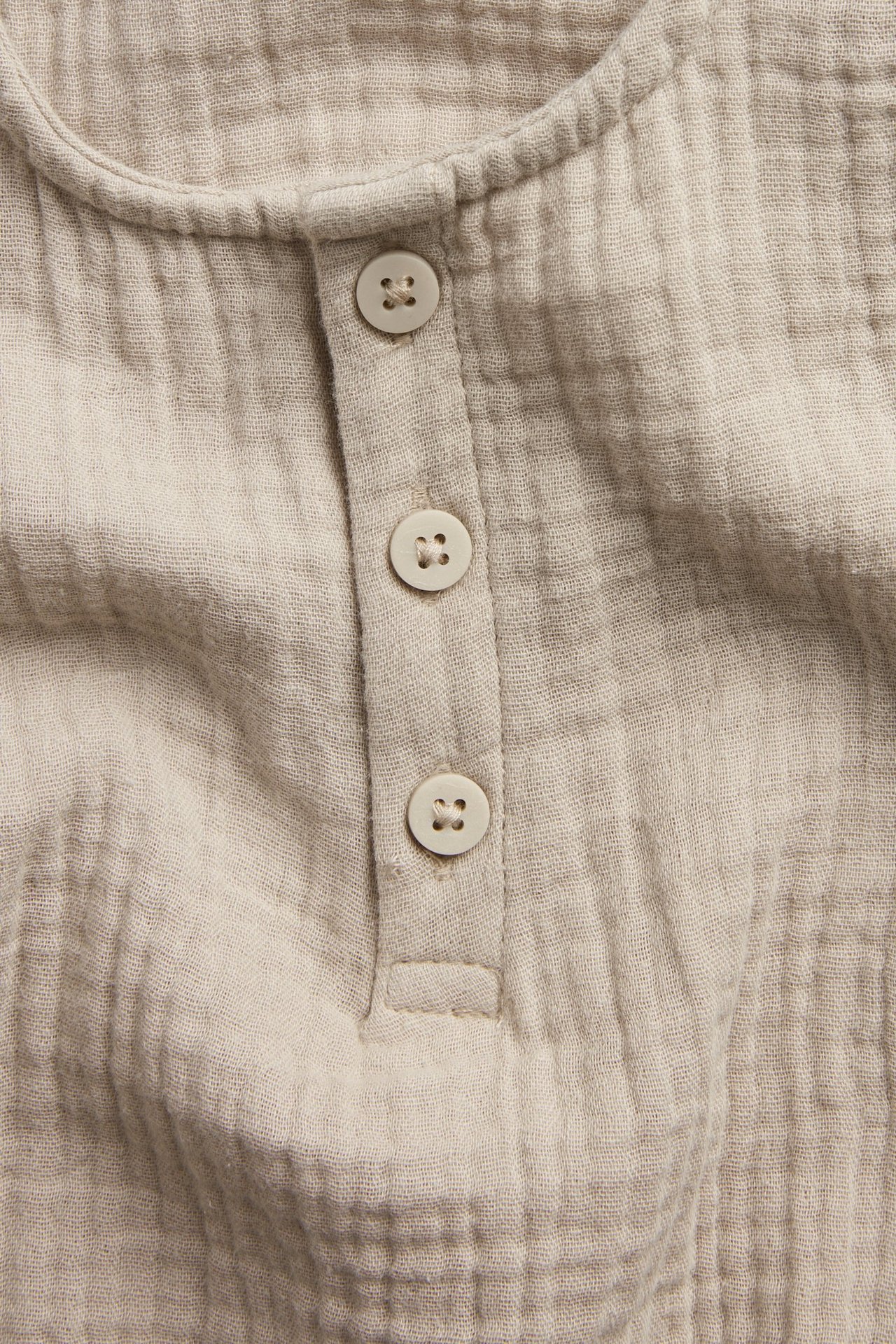Zbliżenie z przodu beżowego body niemowlęcego z muślinu, z trzema jasnymi guzikami.