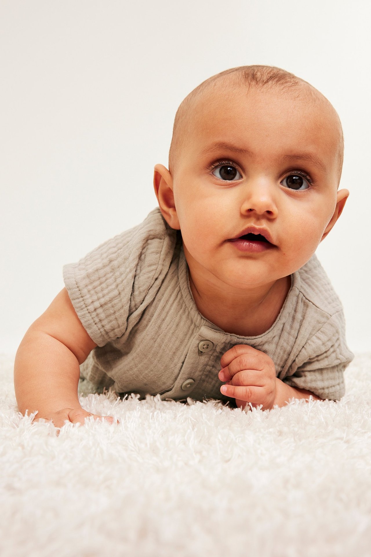 Framifrån, baby i beige kortärmad muslinbody med knappar fram, liggande på en vit fluffig matta.