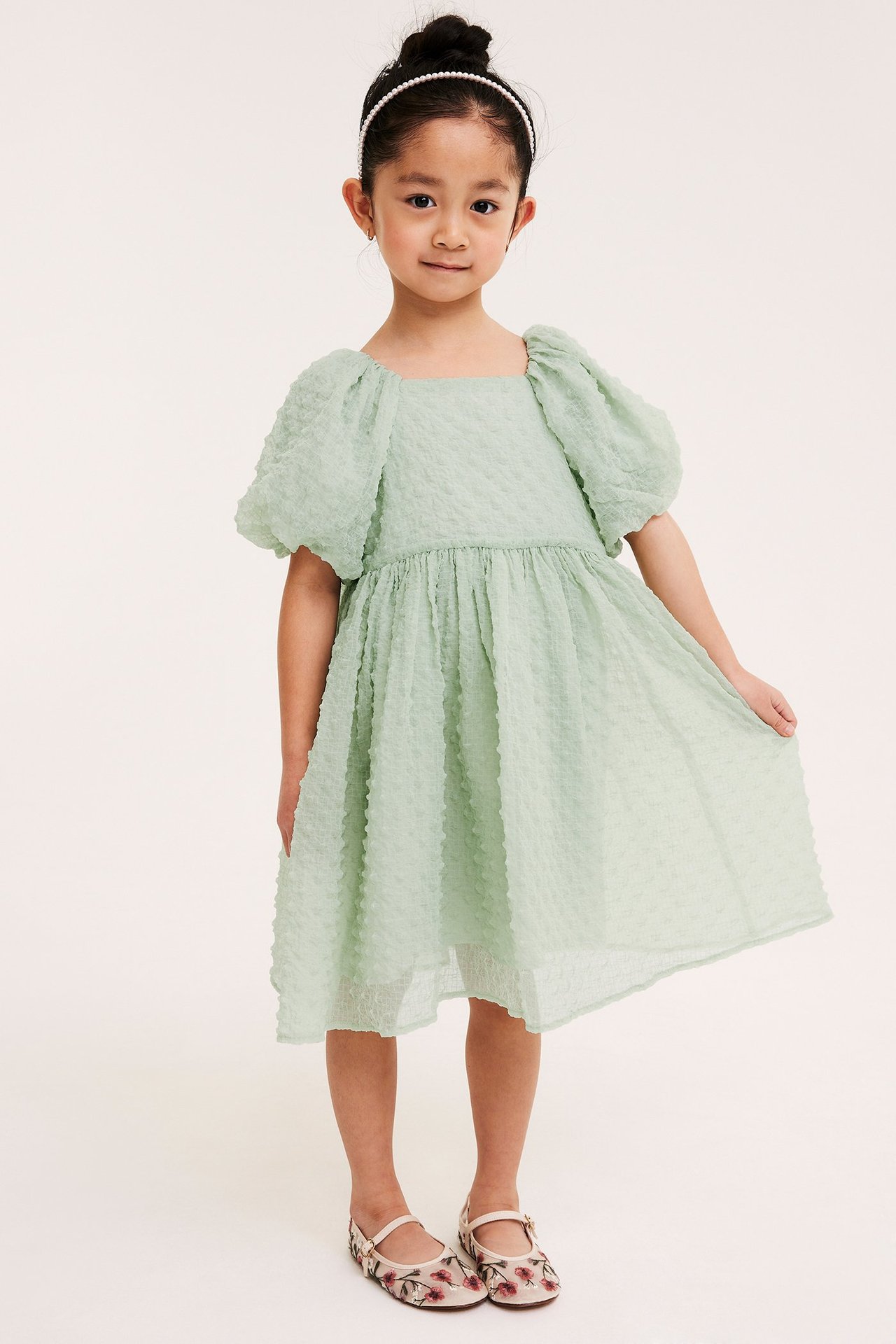 Lapsi edestä yllään runsas vaaleanvihreä kuvioitu sifonkimekko, helmipanta ja kukkakirjaillut matalat kengät.