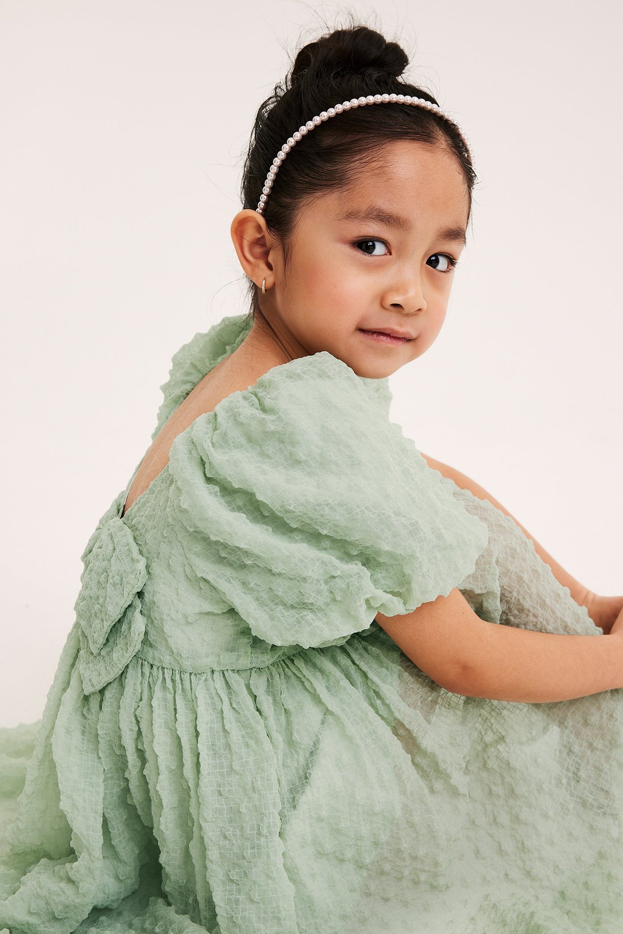 Lapsi vihreässä runsaassa sifonkimekkossa takarusetilla ja helmipannalla, takaviisto näkymä.