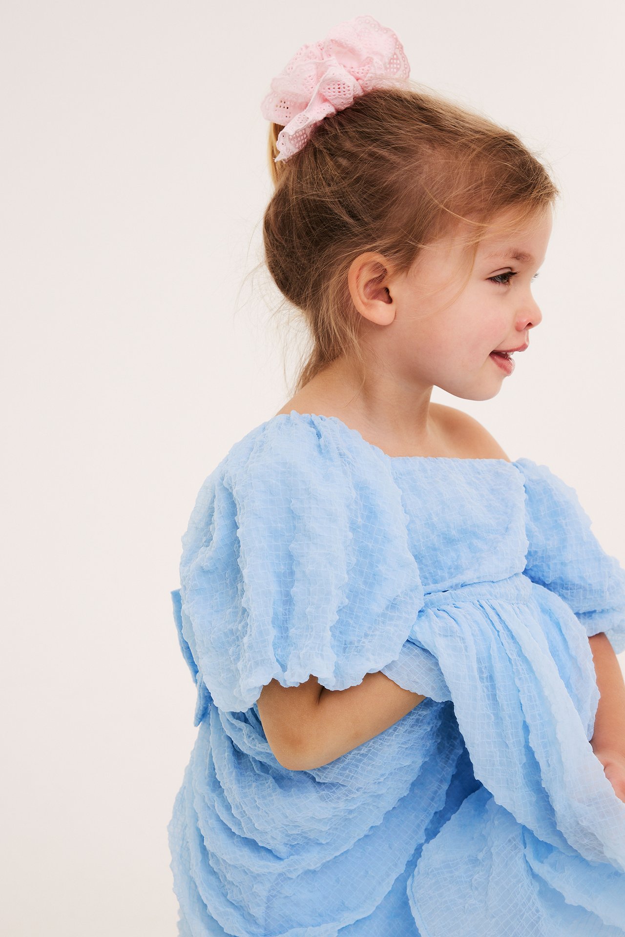 Sidovy av ett barn i en ljusblå texturerad chiffongklänning och en rosa spets-scrunchie.