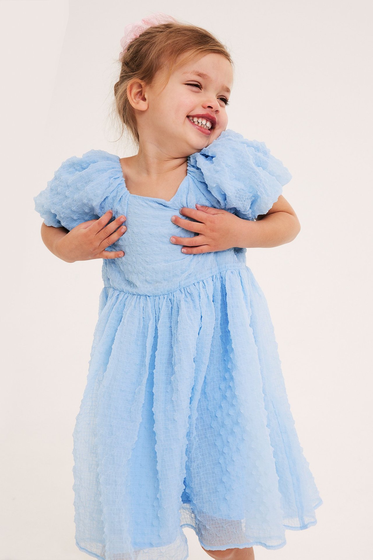Leende barn i en ljusblå chiffongklänning med puffärmar och en rosa håraccessoar, framifrån.