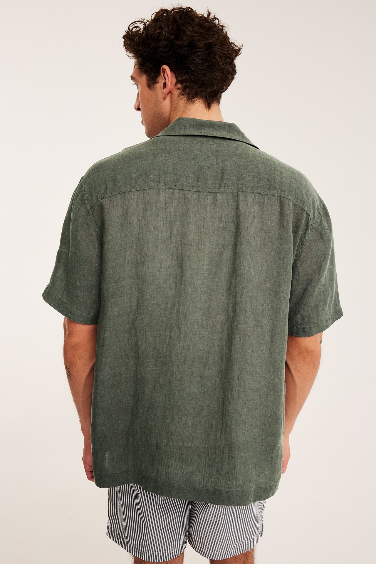 Mies takaa päin vihreässä meleeratussa lyhythihaisessa pellavapaidassa ja raidallisissa shortseissa.