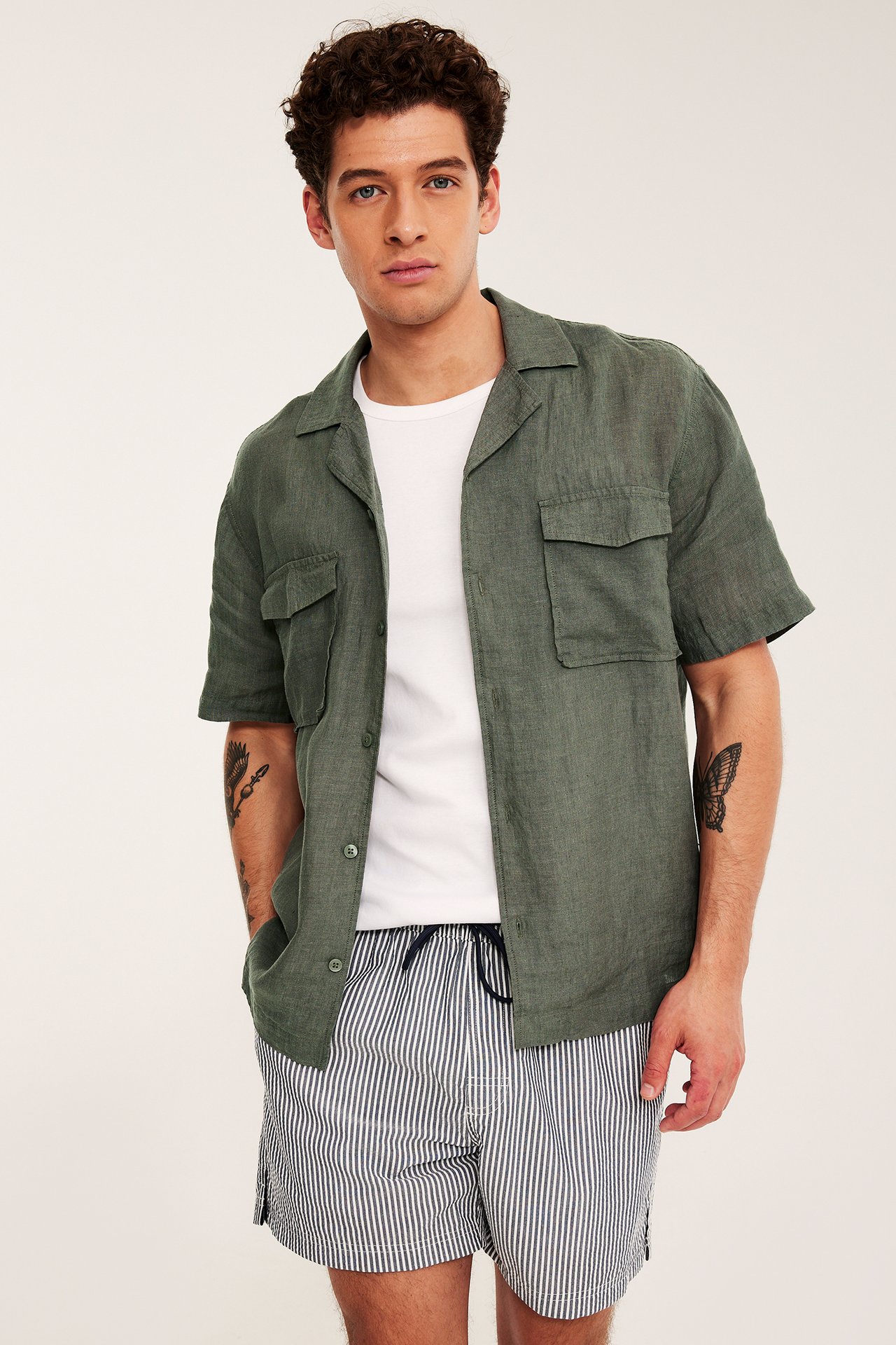 Framifrån: man i öppen melerad grön kortärmad linneskjorta, vit t-shirt och randiga shorts.