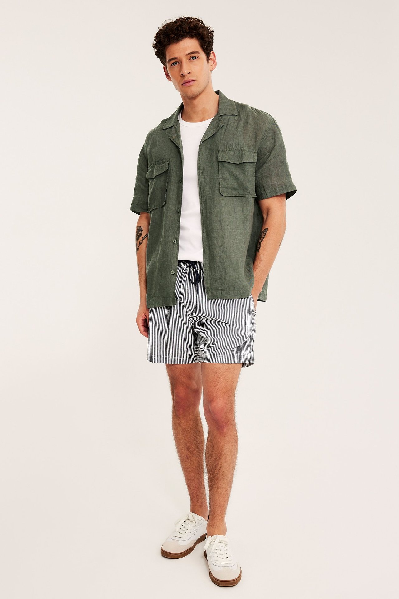 Edestä: Mies vihreä lyhythihainen pellavapaita, valkoinen t-paita, raidalliset shortsit, valkoiset tennarit.