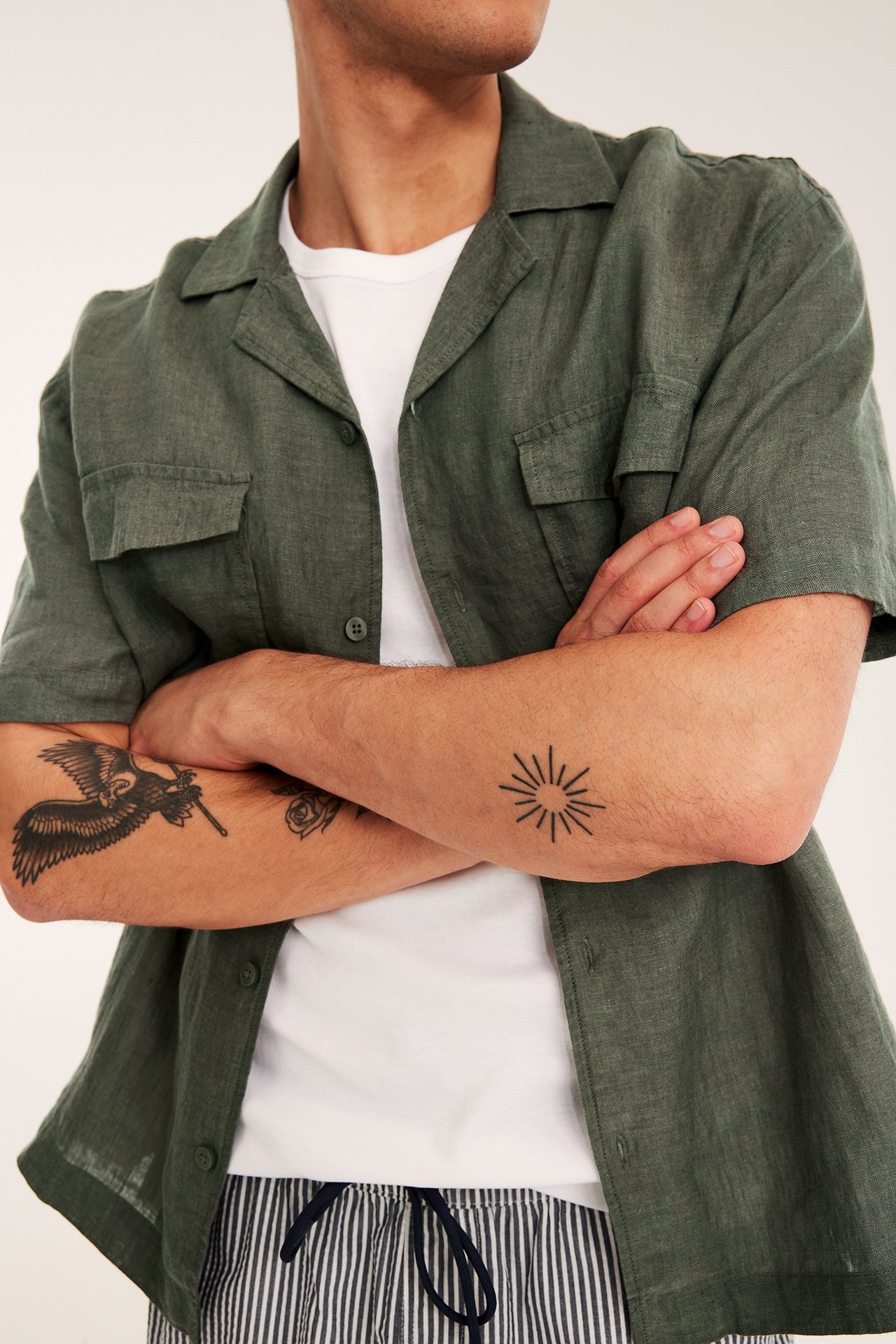 Front view: Man i en öppen grönmelerad kortärmad linneskjorta över vit t-shirt och randiga shorts.