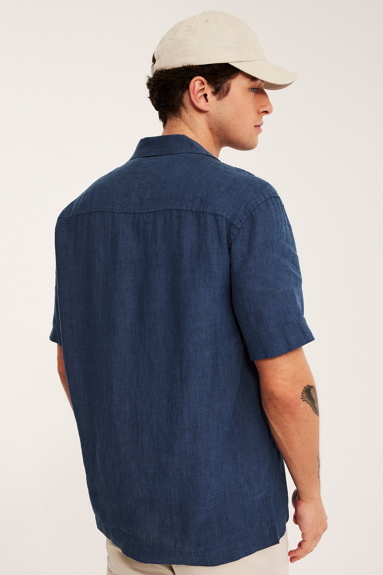 Mies takaa päin sinisessä lyhythihaisessa pellavapaidassa ja vaaleanbeigessä lippalakissa.