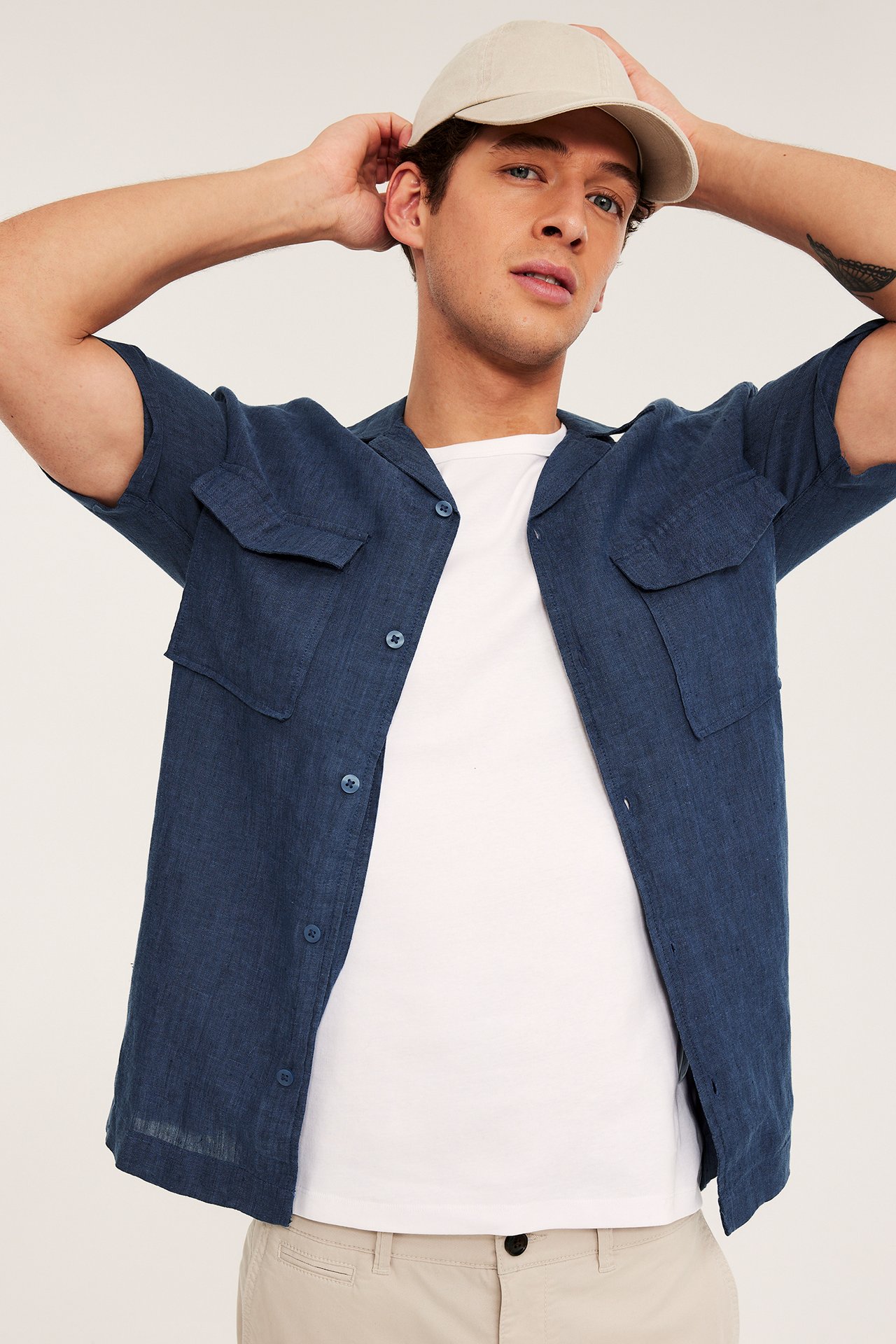Man bär en mörkblå kortärmad linneskjorta med fickor över en vit t-shirt, beige chinos och keps.