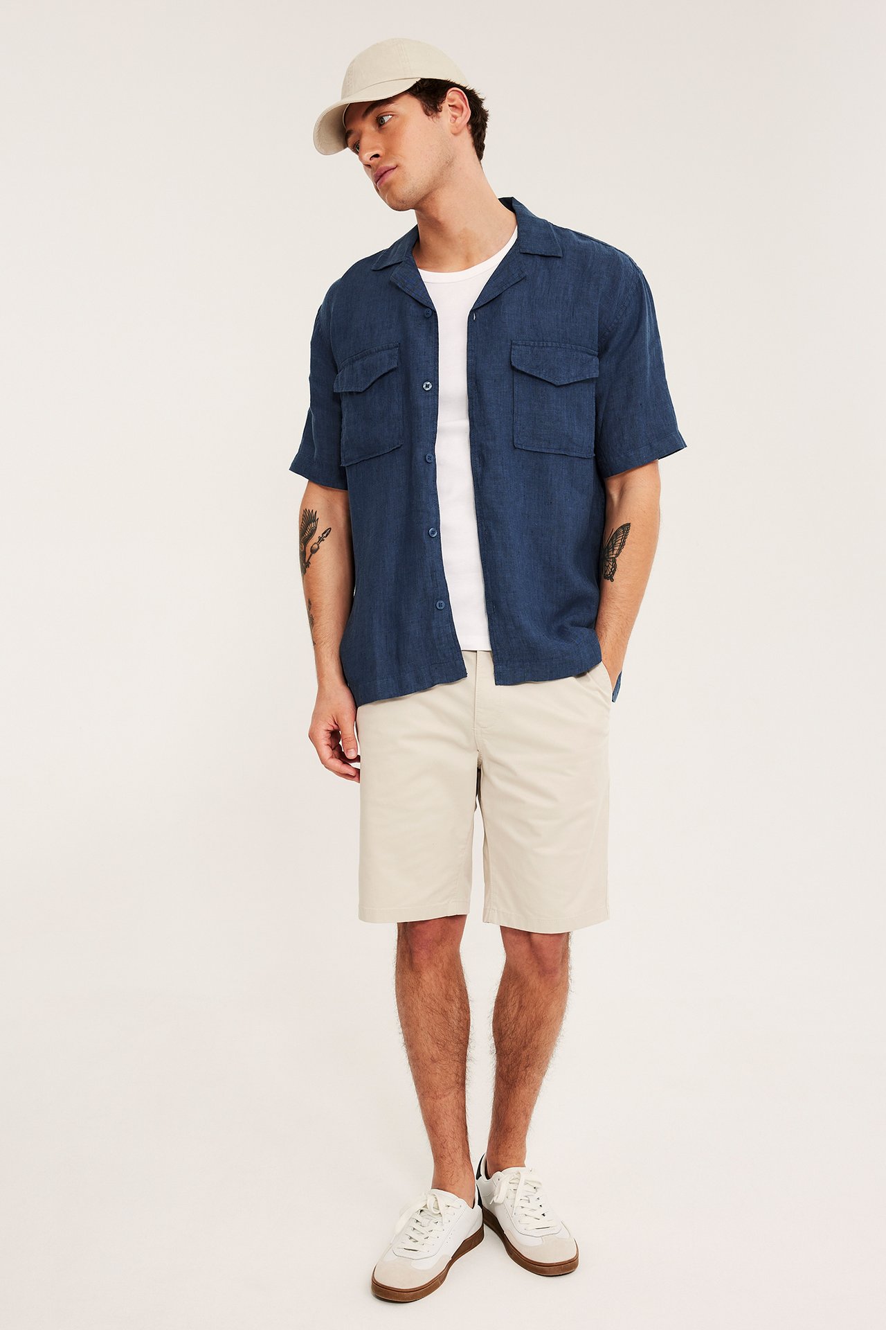 Framifrån: Man bär öppen blåmelerad linneskjorta, vit t-shirt, beige shorts, vita sneakers och beigekeps.