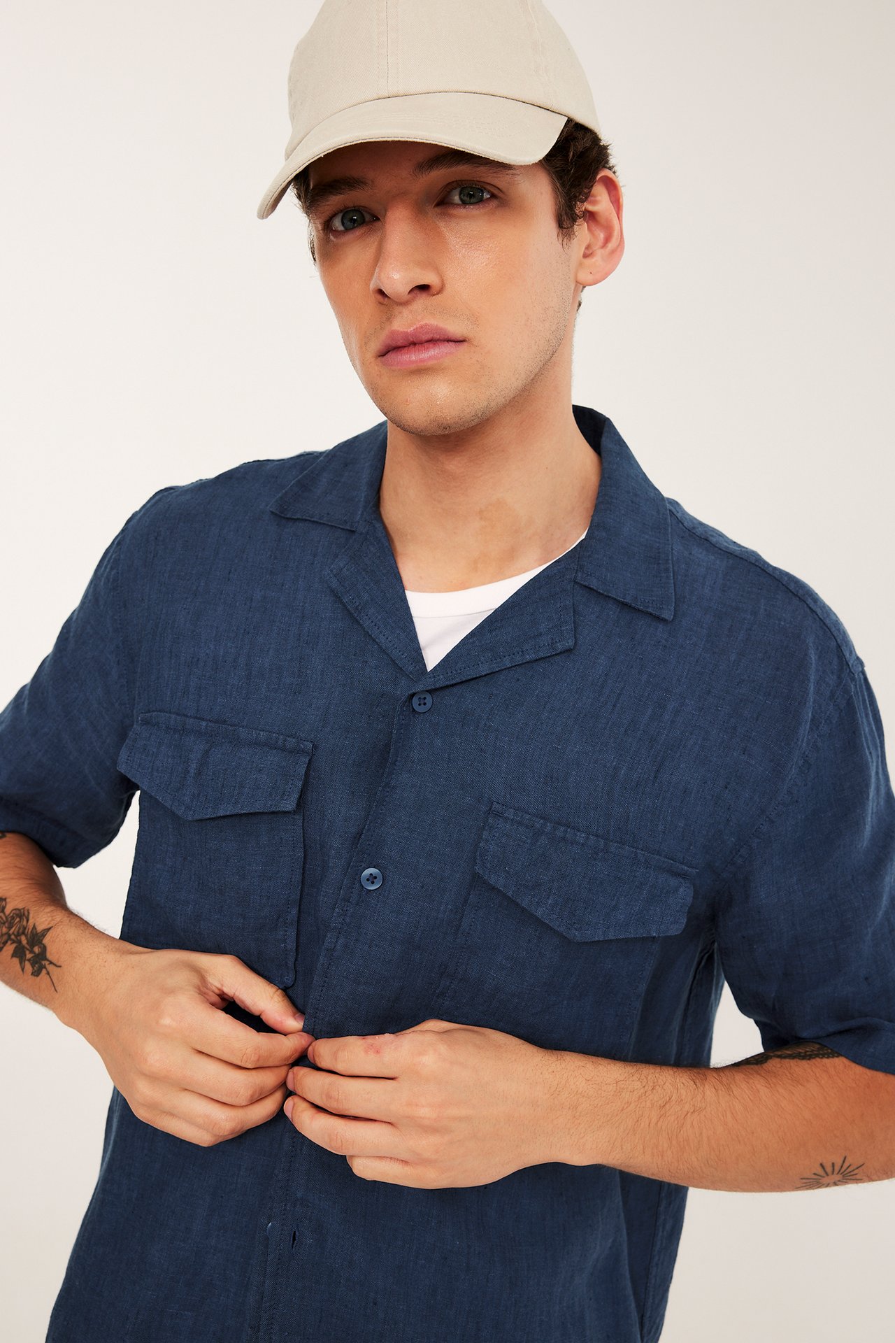 Framifrån: Man knäpper blåmelerad kortärmad linneskjorta med bröstfickor, bär vit t-shirt, beige keps.