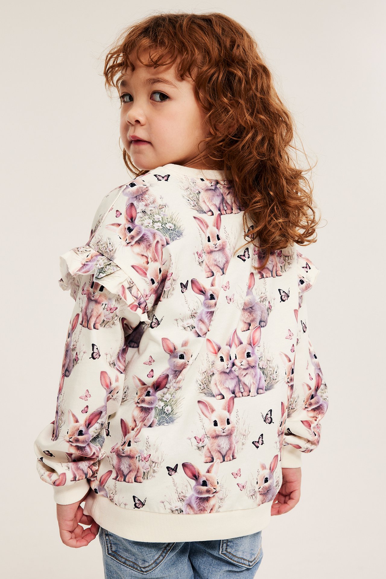 Lapsi takaa päin luonnonvalkoisessa puuvillapaidassa, jossa kanikuvio ja röyhelöhihat.