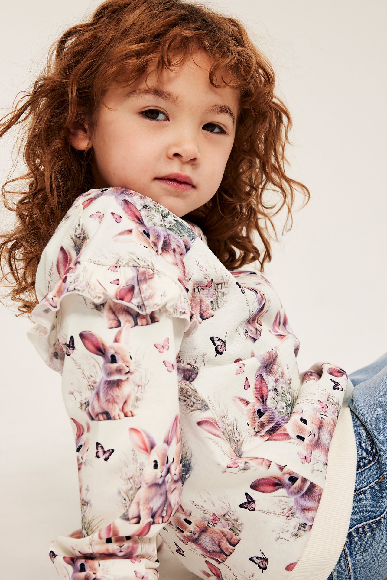 Närbild på ett barn i offwhite bomullströja med kaninmönster, volangärmar och blå jeans.