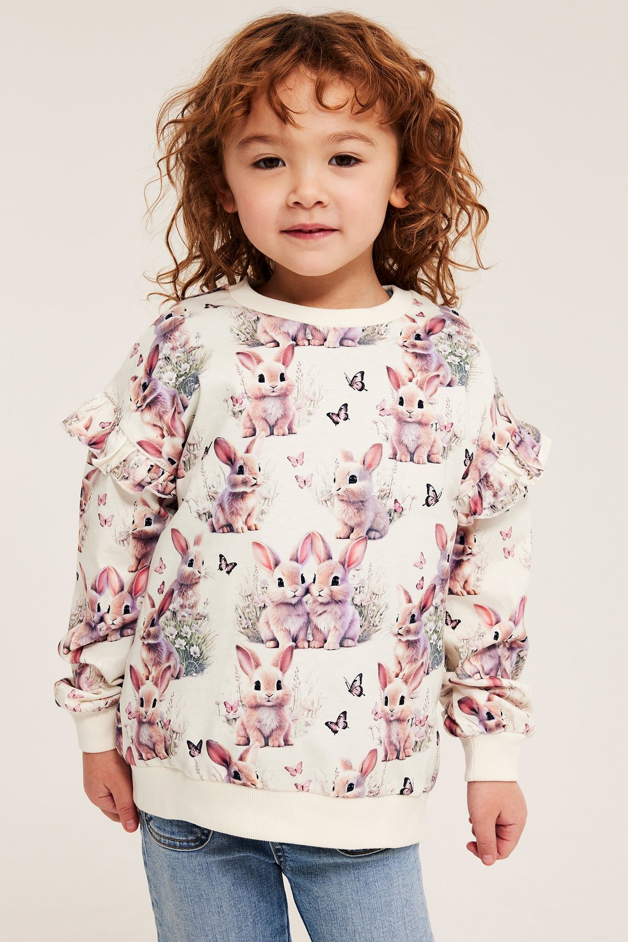 Lapsi edestä, offwhite puuvillapaidassa, jossa röyhelöhihat ja jänismalli, sinisten farkkujen päällä.