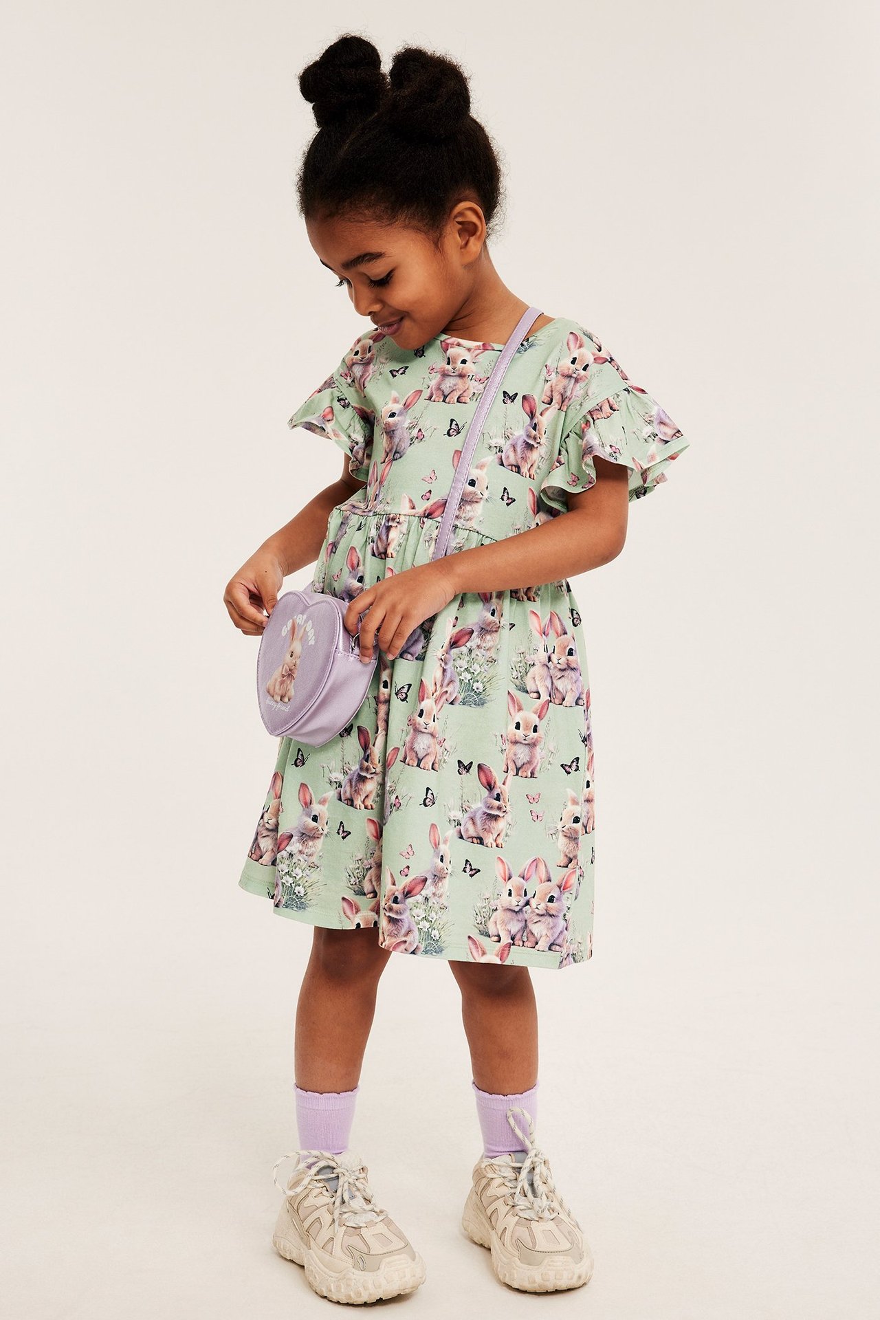 Widok z przodu: dziecko w zielonej bawełnianej sukience w króliki, z falbankami i fioletową torebką.