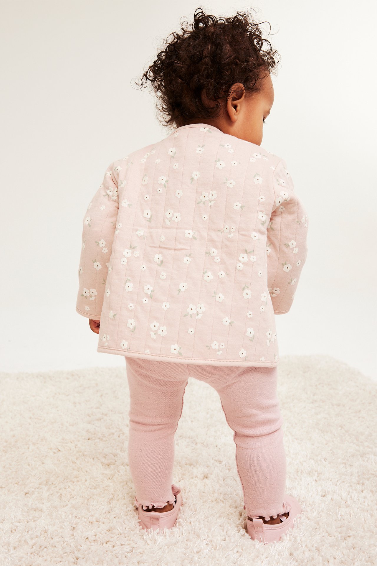 Widok z tyłu: dziecko w różowej kurtce pikowanej w kwiaty, różowych legginsach i bucikach z falbanką.
