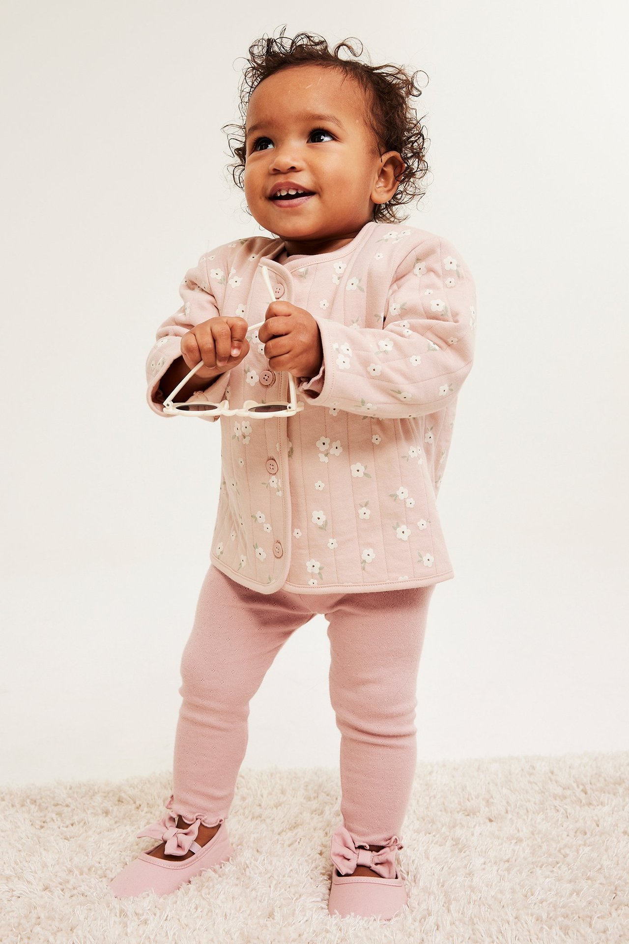 Etunäkymä: vauva vaaleanpunaisessa tikatussa kukkajakussa, leggingseissä, rusettikengissä ja pitää valkoisia aurinkolaseja.