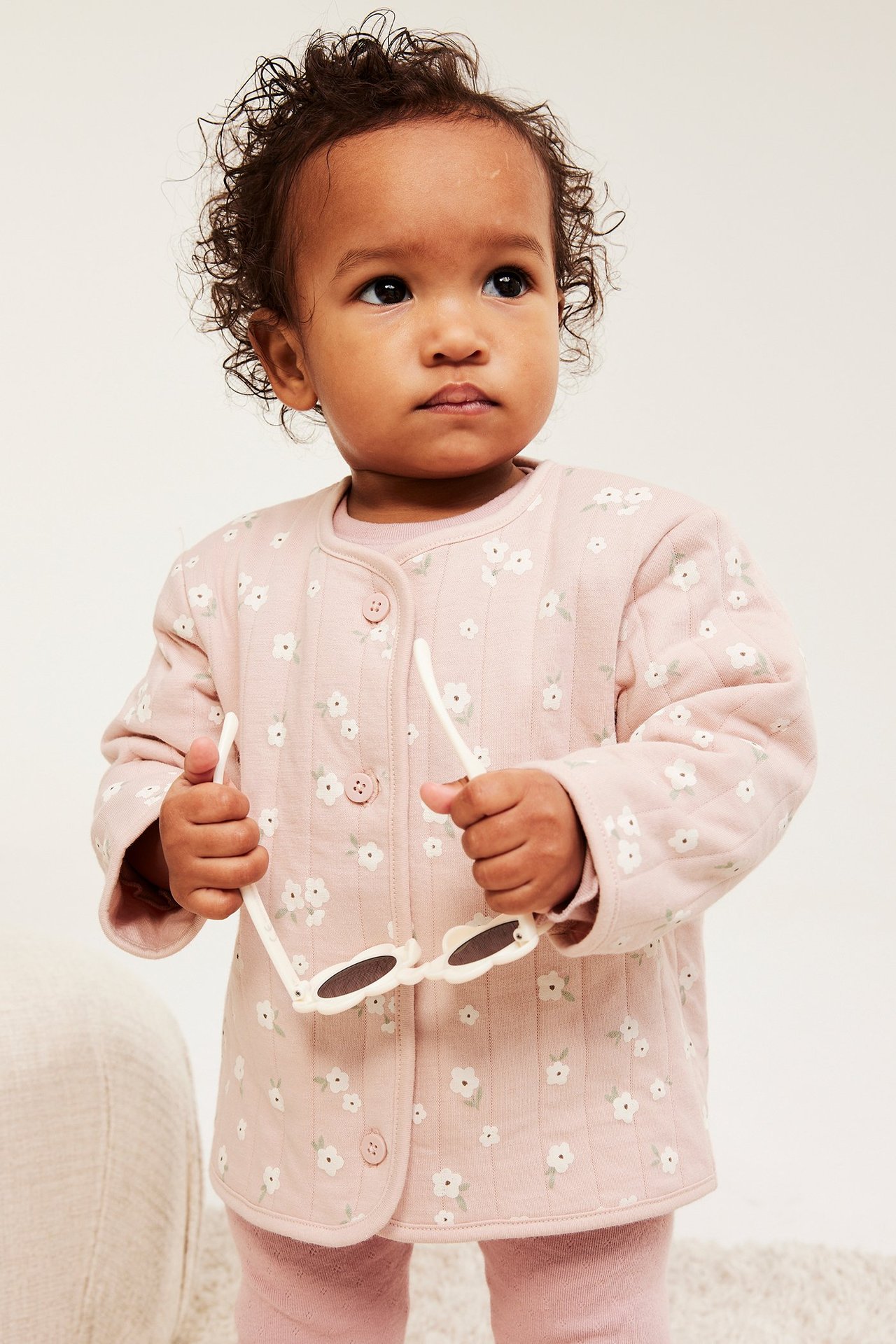 Widok z przodu: Dziecko w różowej pikowanej kurtce w kwiaty na guziki, różowym topie, legginsach, trzyma białe okulary.