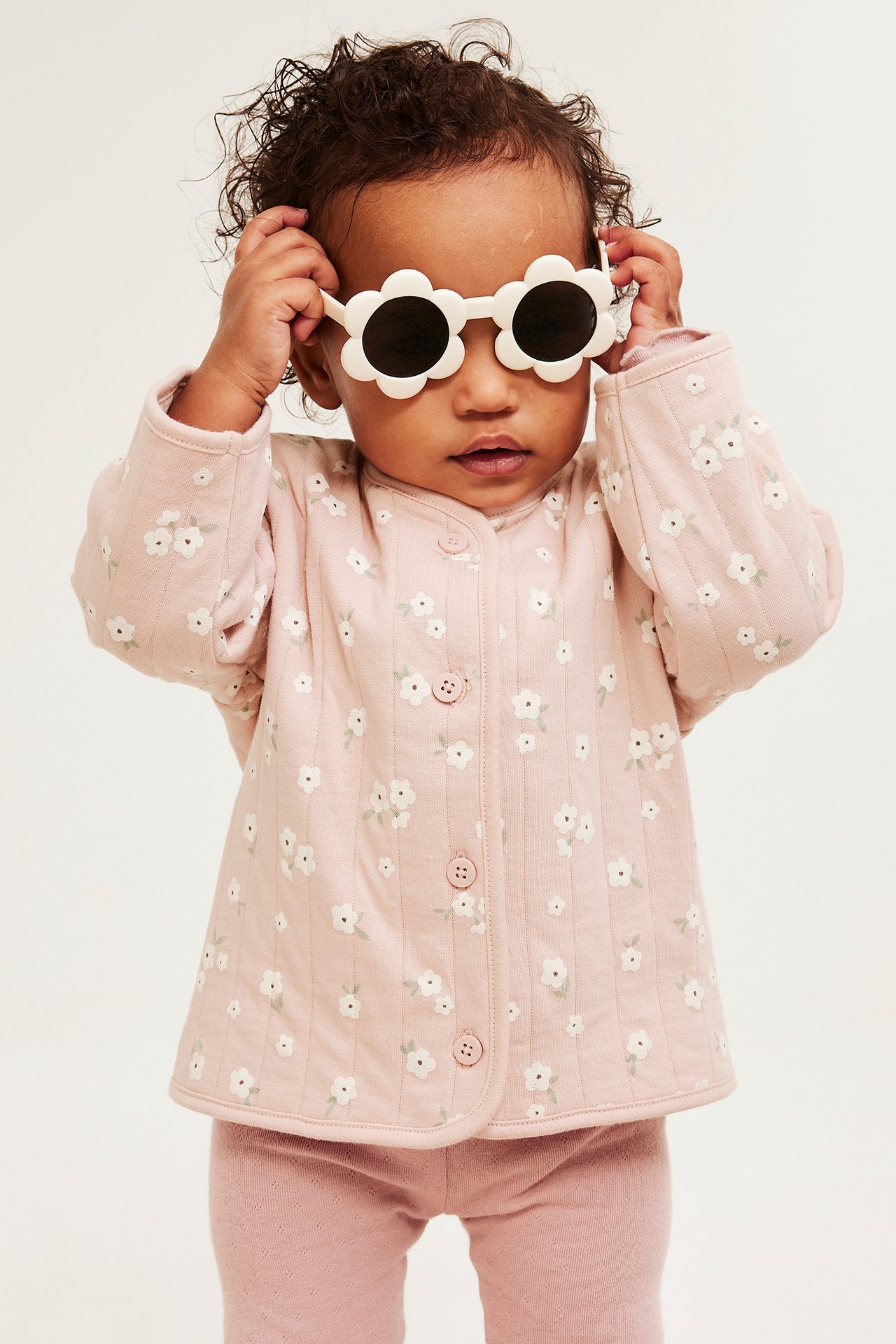 Niemowlę w różowej pikowanej kurtce z białym kwiatowym nadrukiem, guzikami i białych okularach-kwiatach.