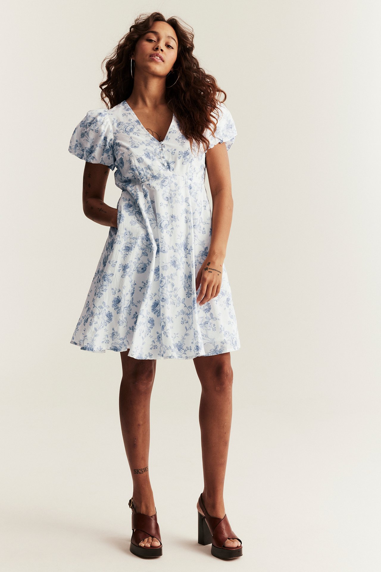 Nainen luonnonvalkoisessa kukkakuosisessa puuvillamekossa puffihihoilla, ruskeat sandaalit ja vannekorvakorut.