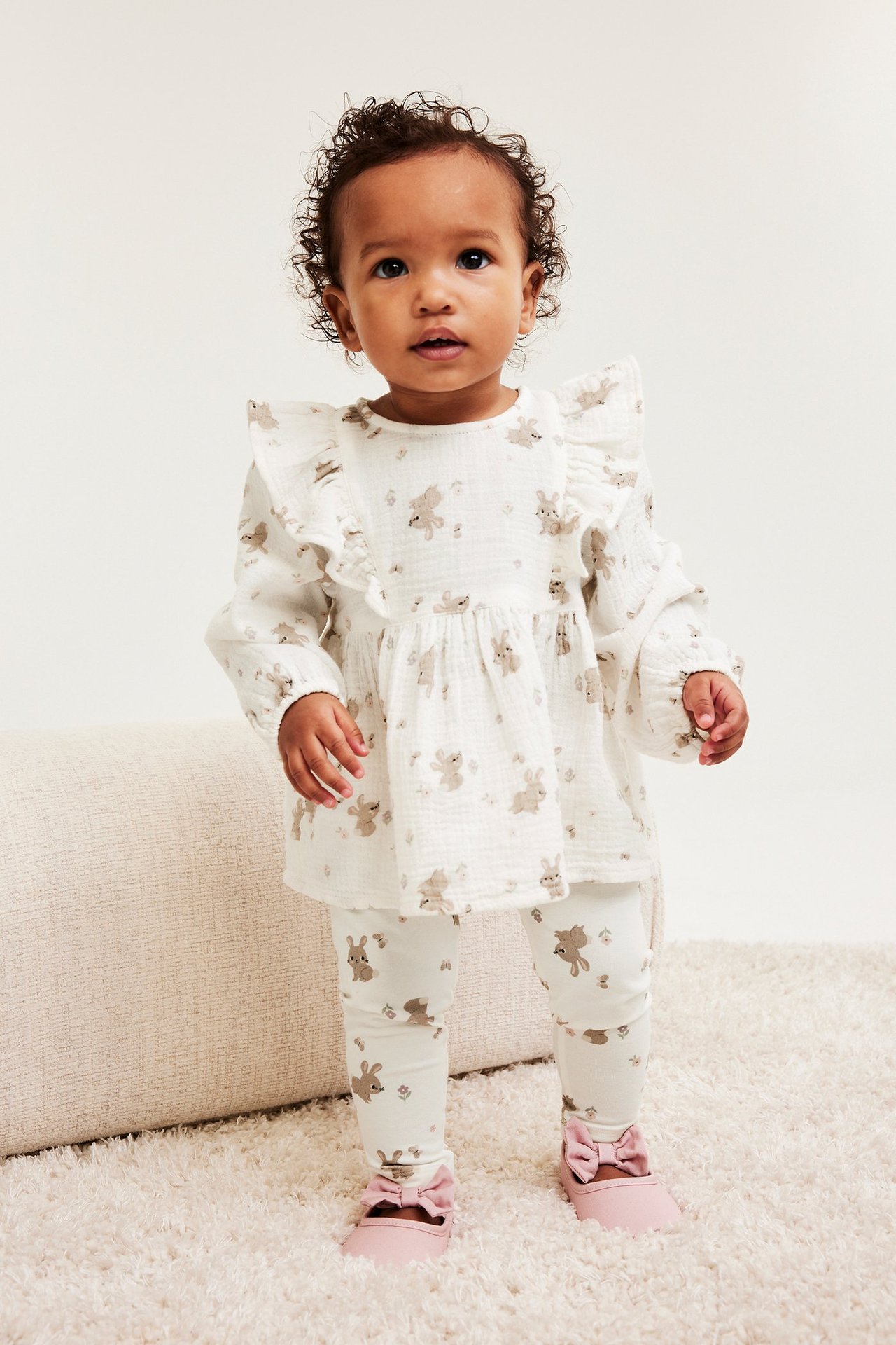 Framifrån: Baby i offwhite muslinblus med volanger, kaninmönster, matchande leggings och rosa rosettskor.