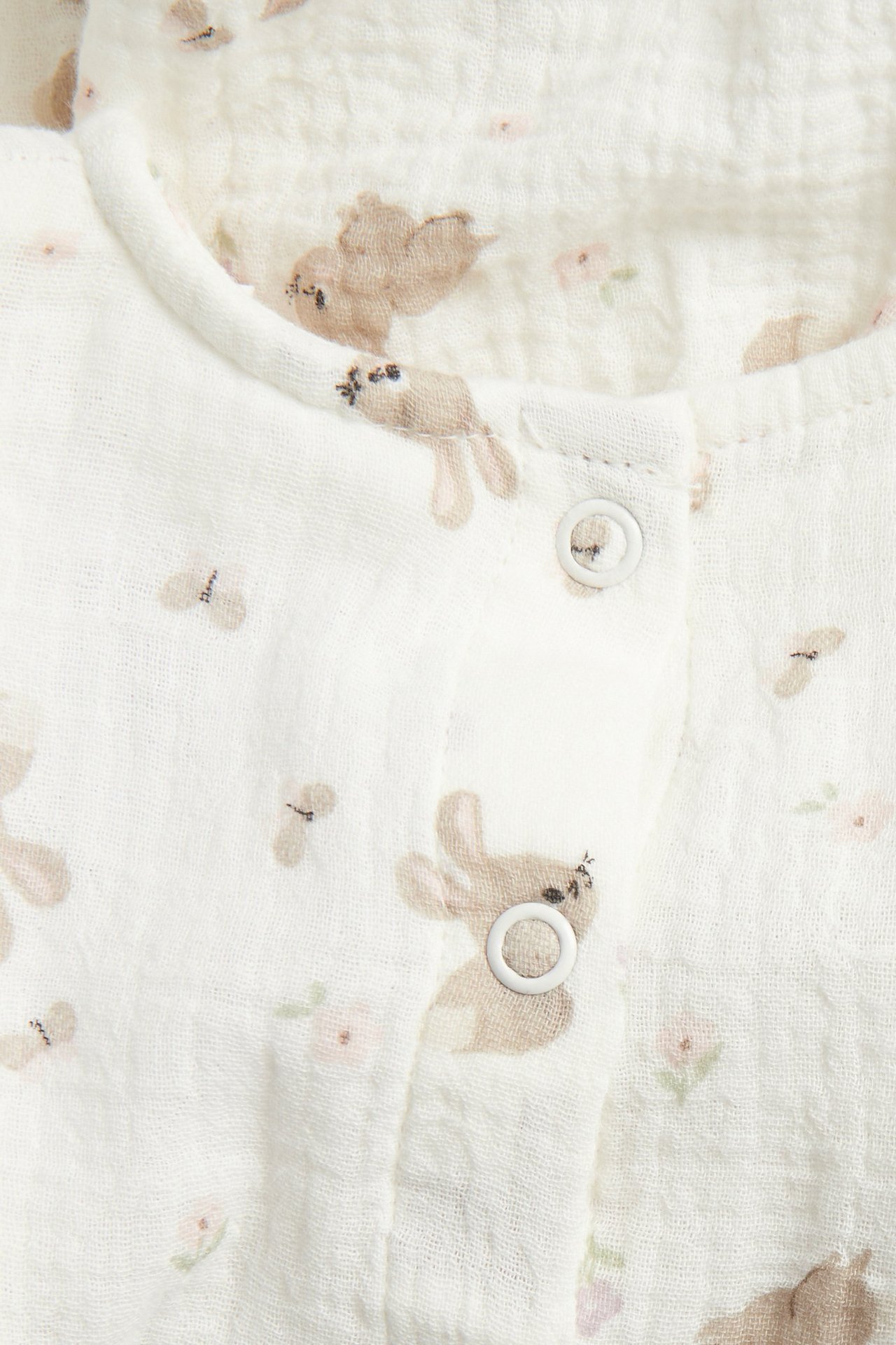 Nærbilde av en offwhite babybluse i bomullsmuslin med trykte brune kaniner og hvite trykknapper.