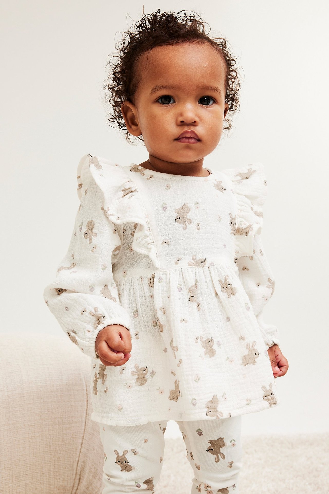 Widok z przodu: Niemowlę w ecru bluzce muślinowej z falbanami i wzorem królików oraz pasujących legginsach.