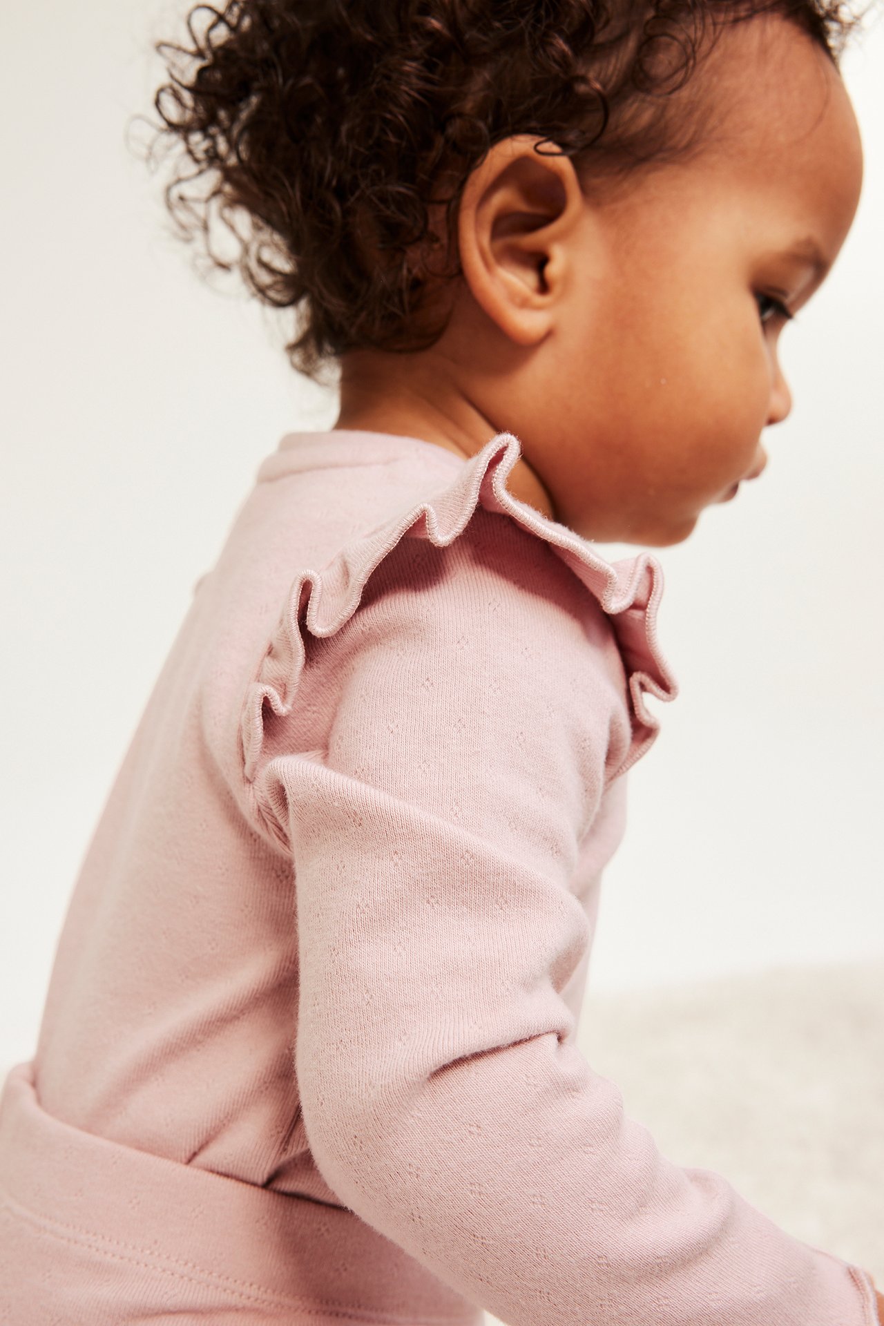 Lähikuva sivulta vauvasta, jolla on vaaleanpunainen pitkähihainen pointelle-body röyhelöillä olkapäillä.