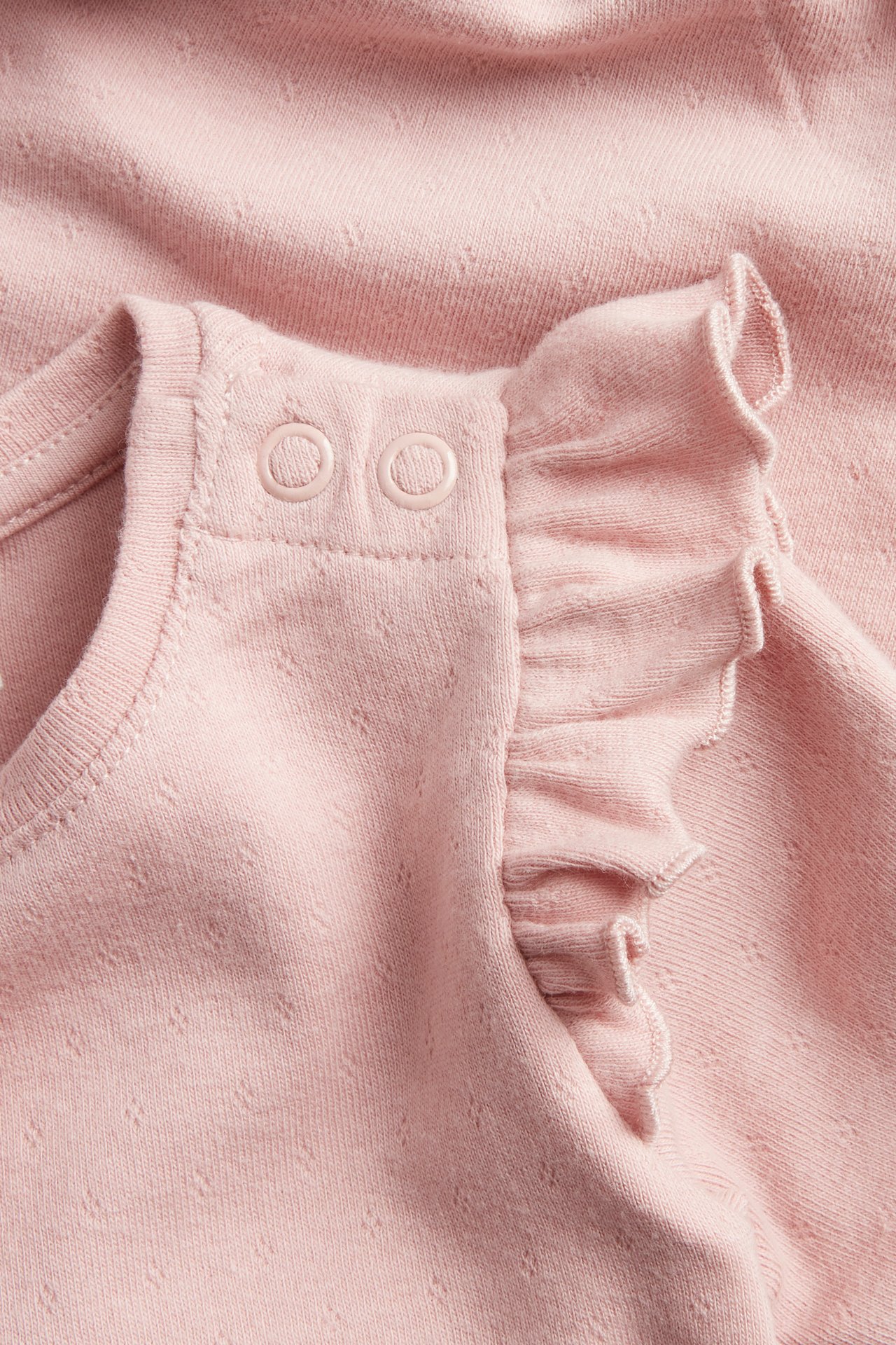 Lähikuva vaaleanpunaisesta pointelle-vauvanbodysta, jossa olkapäällä nepparit ja hihansuissa frillat.