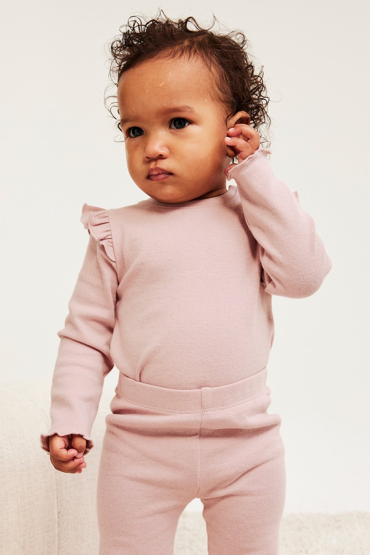 Widok z przodu, niemowlę w różowym body z długim rękawem z falbankami i dopasowanych legginsach.